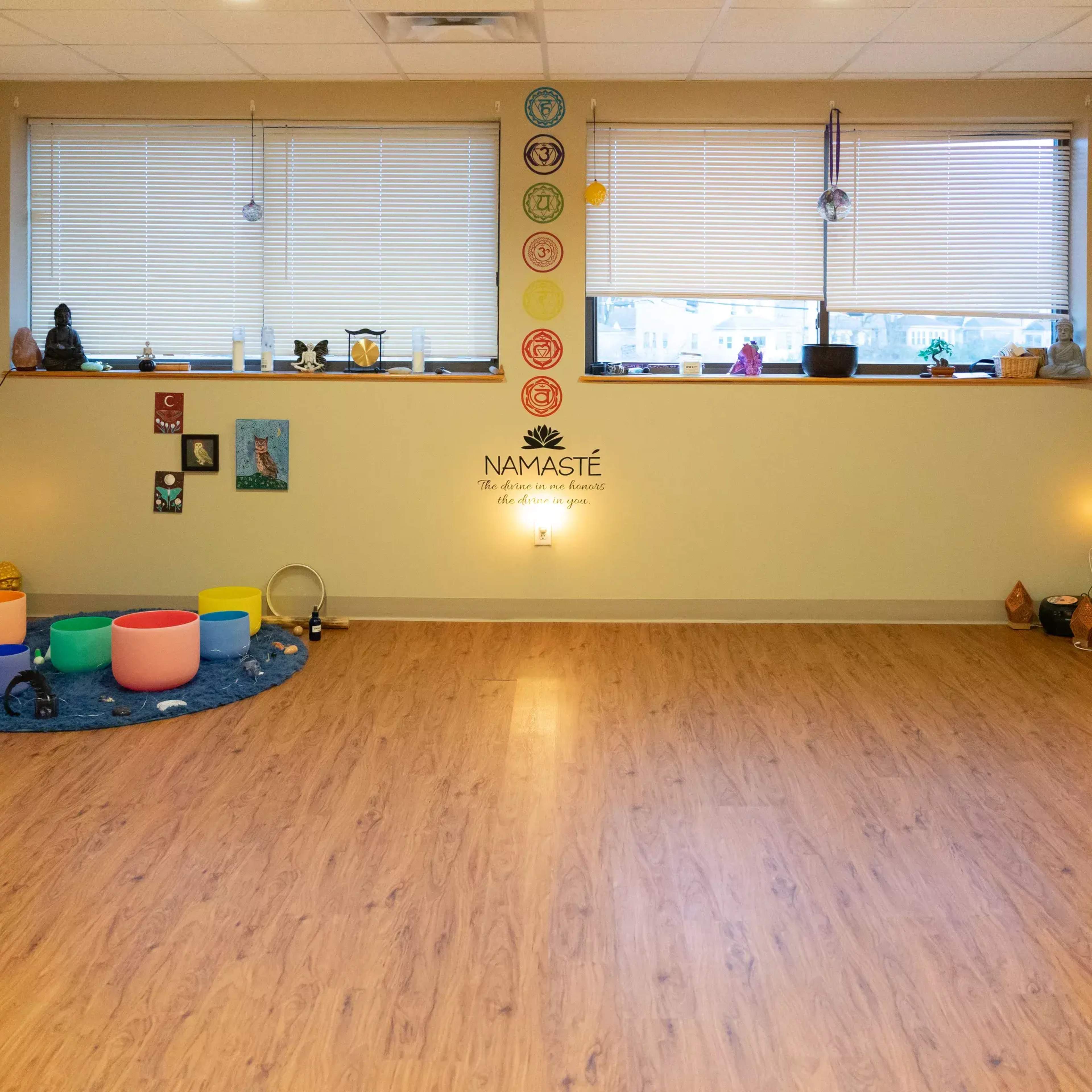 A spacious room features colorful meditation cushions, a circular arrangement of bowls, and decorative items including statues and chakra symbols on the walls.