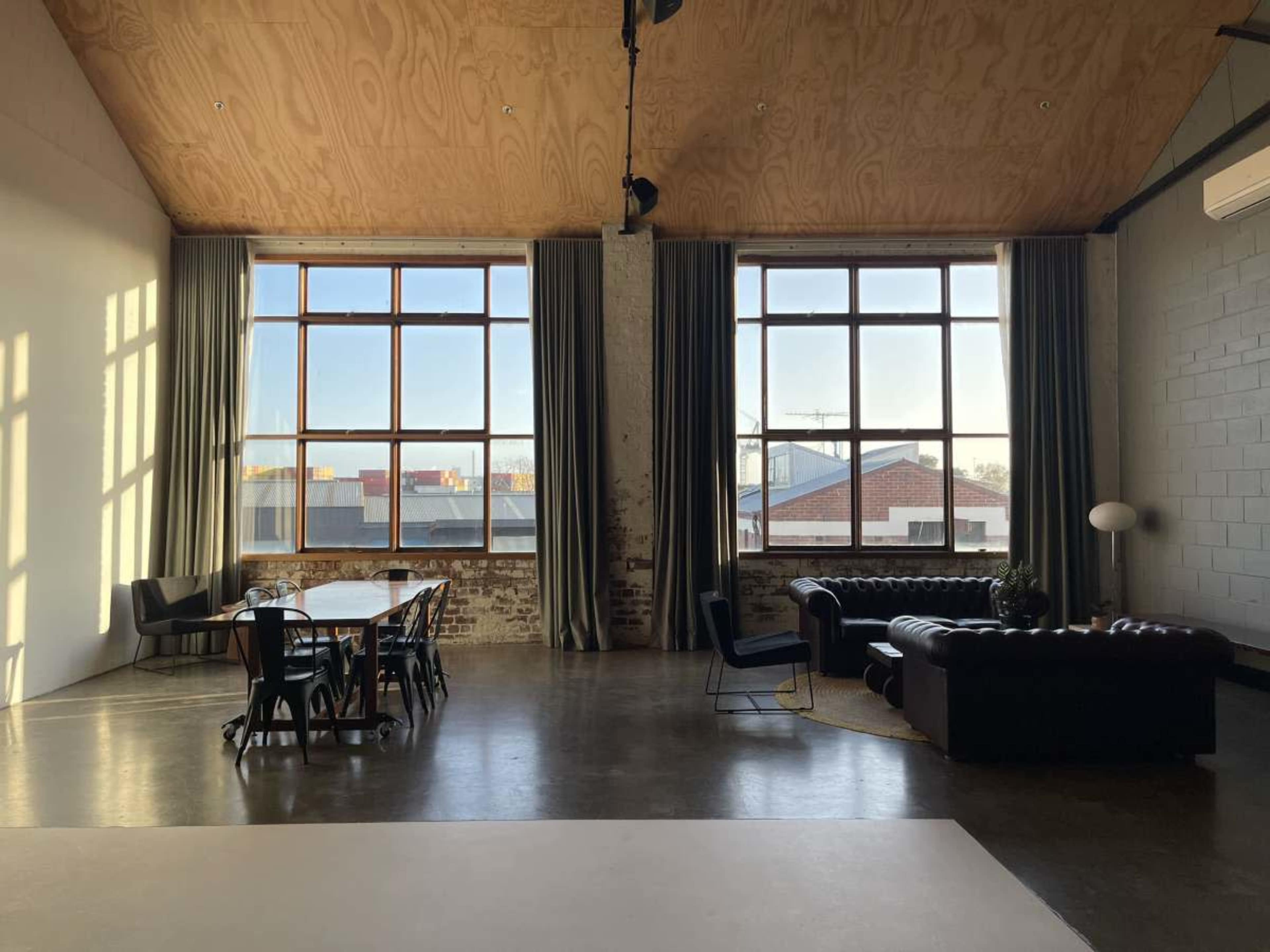 The image shows an industrial-style interior with large windows, a wooden ceiling, a dining table, and a seating area that includes a leather sofa and armchair.