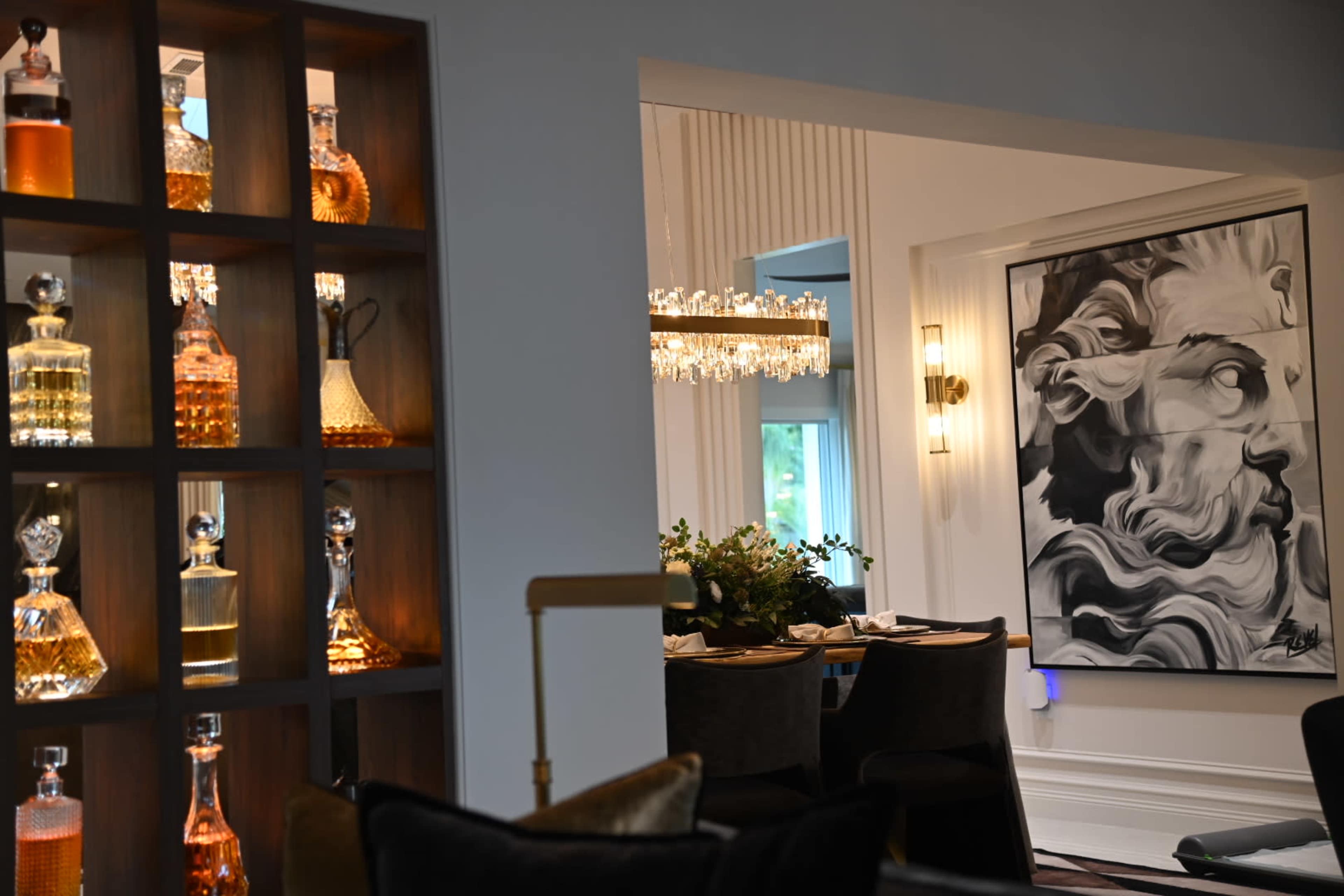 The image features a stylish interior with a backlit display of decorative bottles on one side and an artistic painting of a face on the wall opposite a dining area with a chandelier above the table.