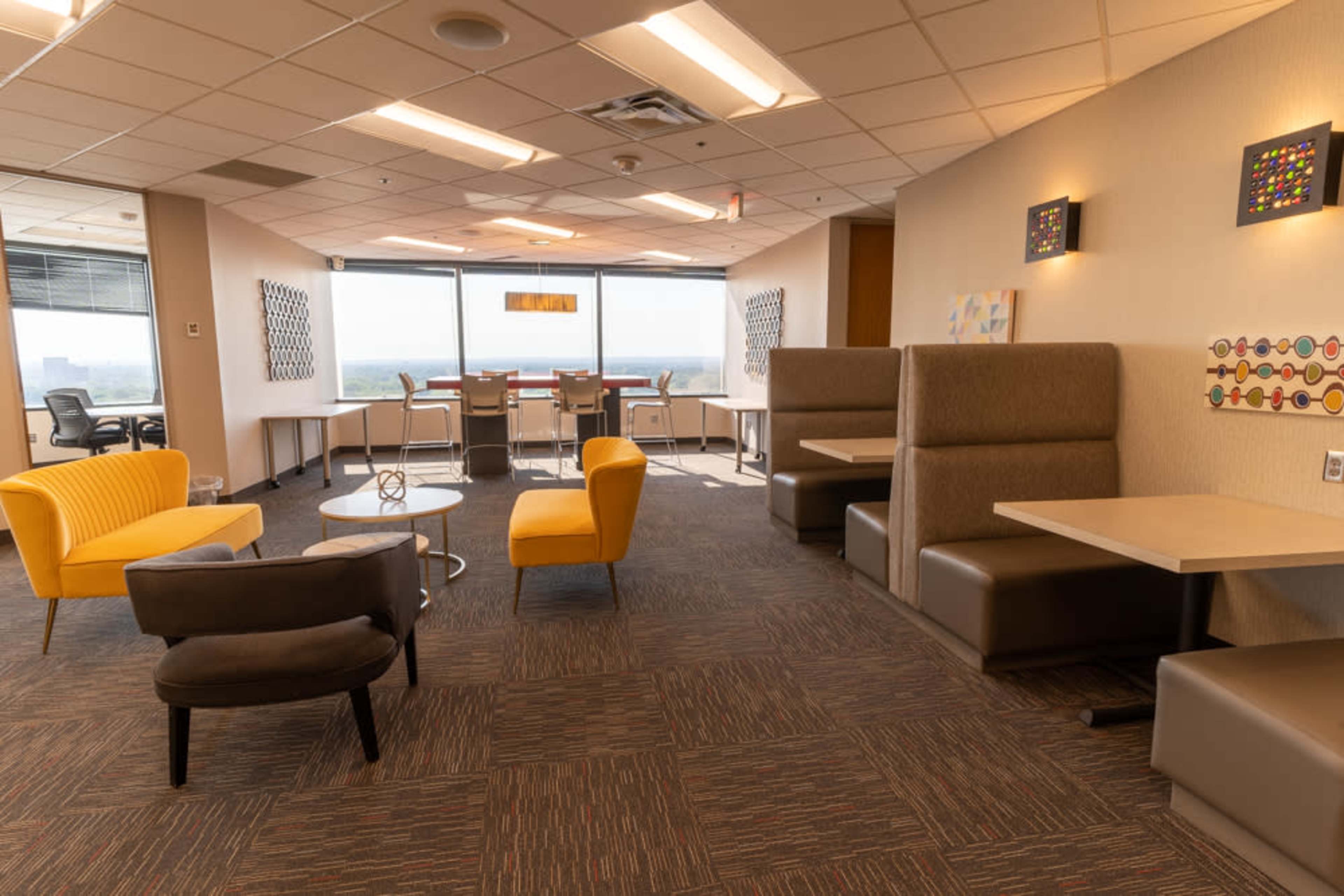 The image shows a modern office lounge area with seating arrangements including yellow and gray chairs, small tables, and large windows providing natural light.