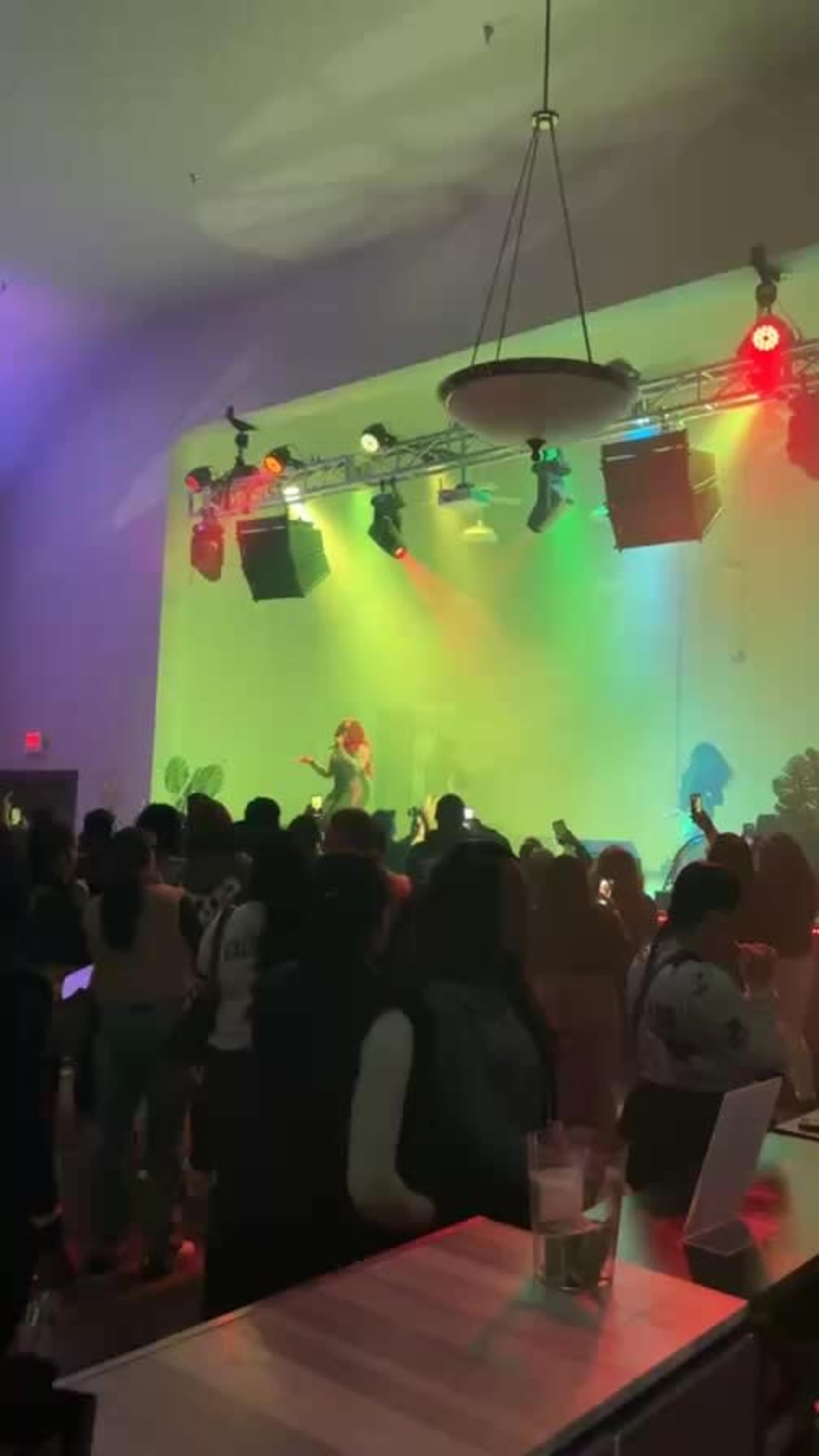 A large crowd gathers in a brightly lit venue while a performer sings on stage, with colorful lights illuminating the scene.