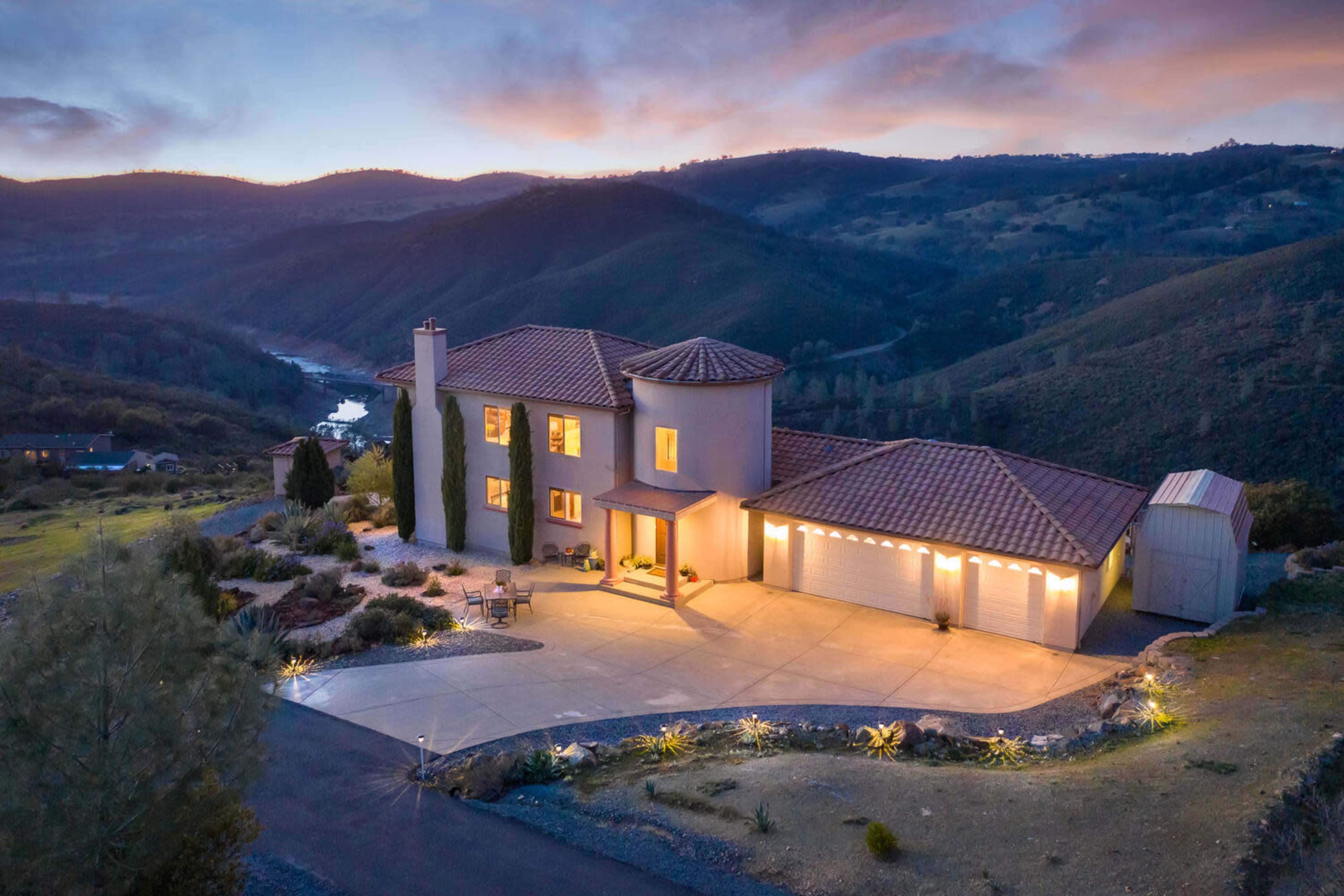 A modern two-story house with a tile roof, surrounded by rolling hills and a river, illuminated by outdoor lighting at dusk.