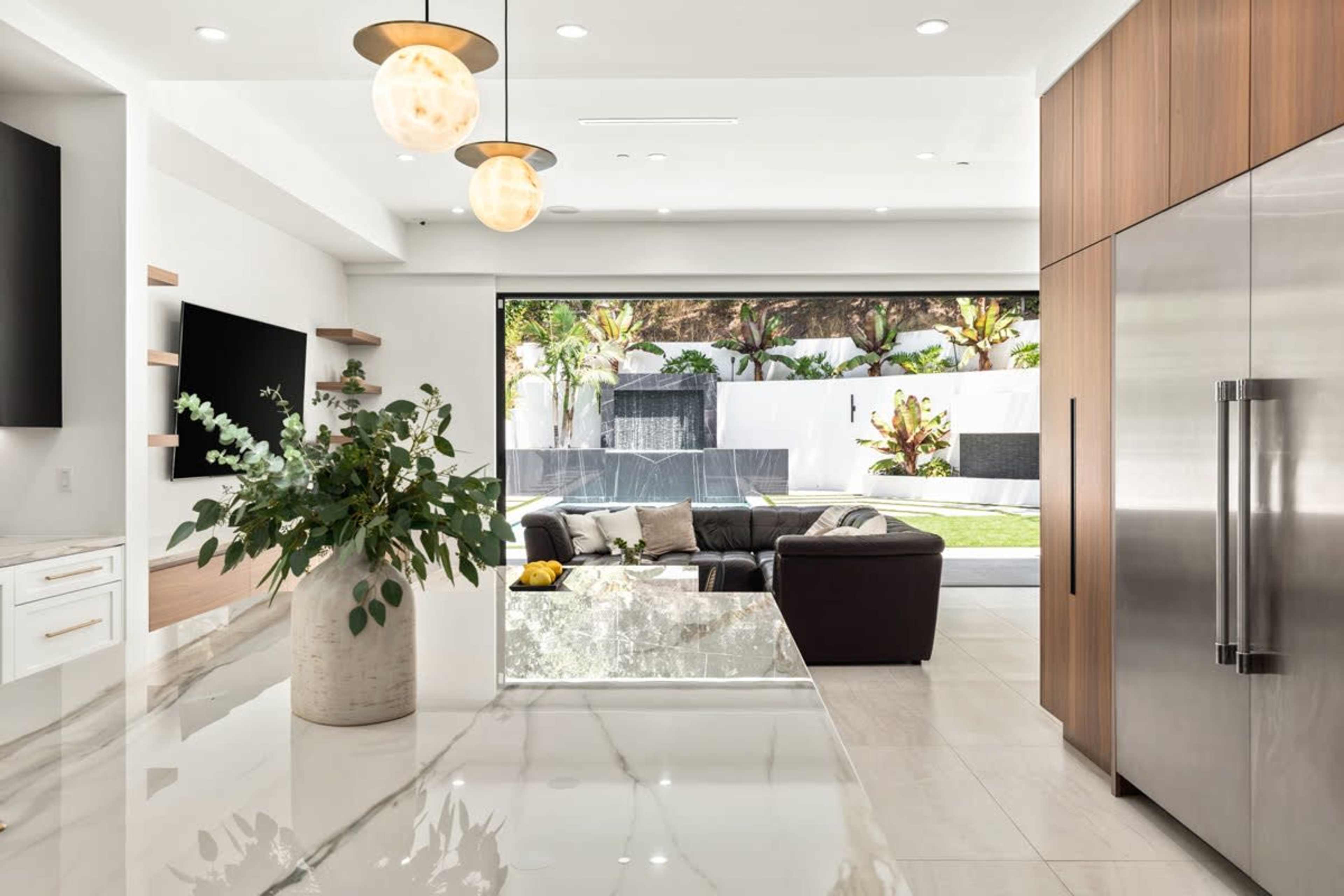 A modern kitchen opens to a backyard, featuring a large island, stainless steel refrigerator, and a dark leather sofa facing large glass doors.