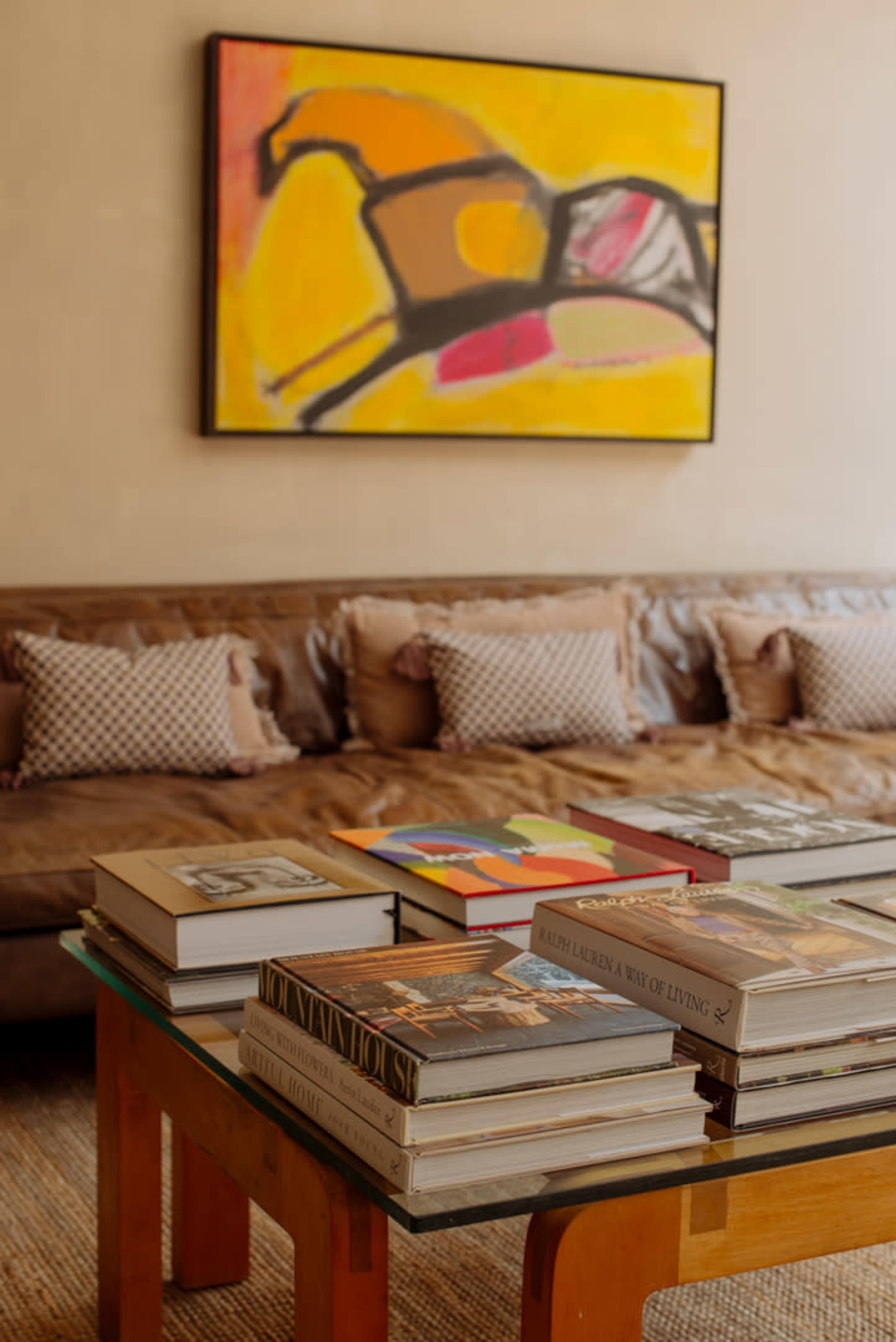 A brown leather sofa sits against a wall adorned with a colorful horse painting, while a glass table displays a stack of various books.