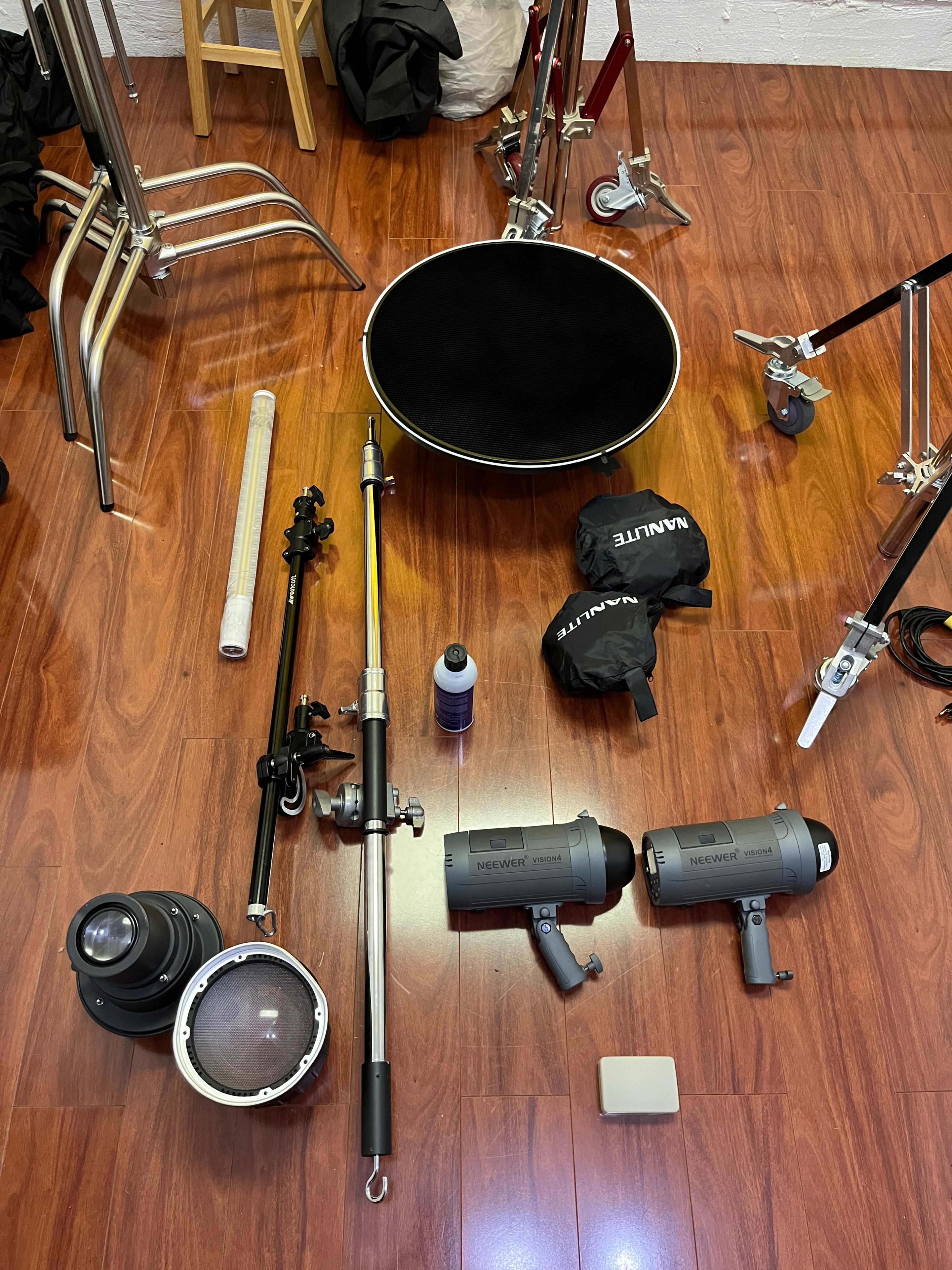 Various photography equipment laid out on a wooden floor, including light stands, reflectors, a lens, and studio lights.