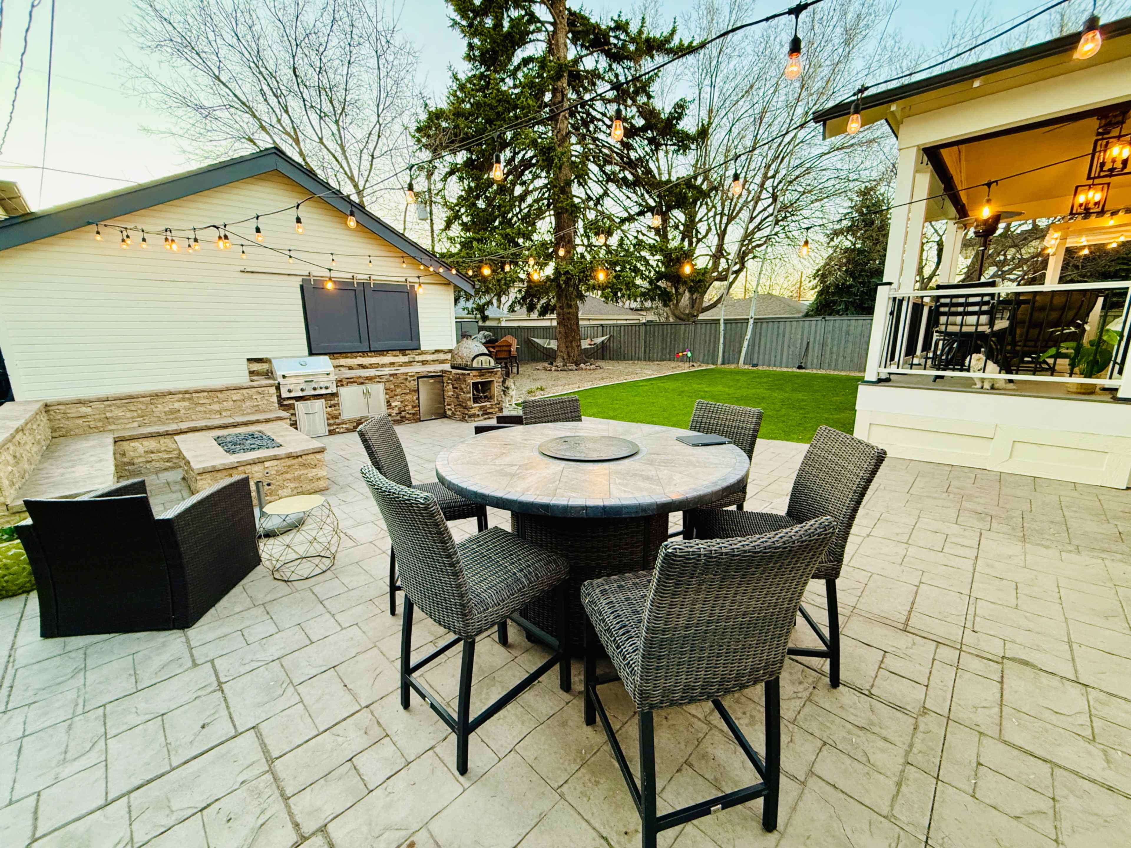 A circular dining table with chairs is set in a backyard featuring string lights, a barbecue area, and a well-manicured lawn.