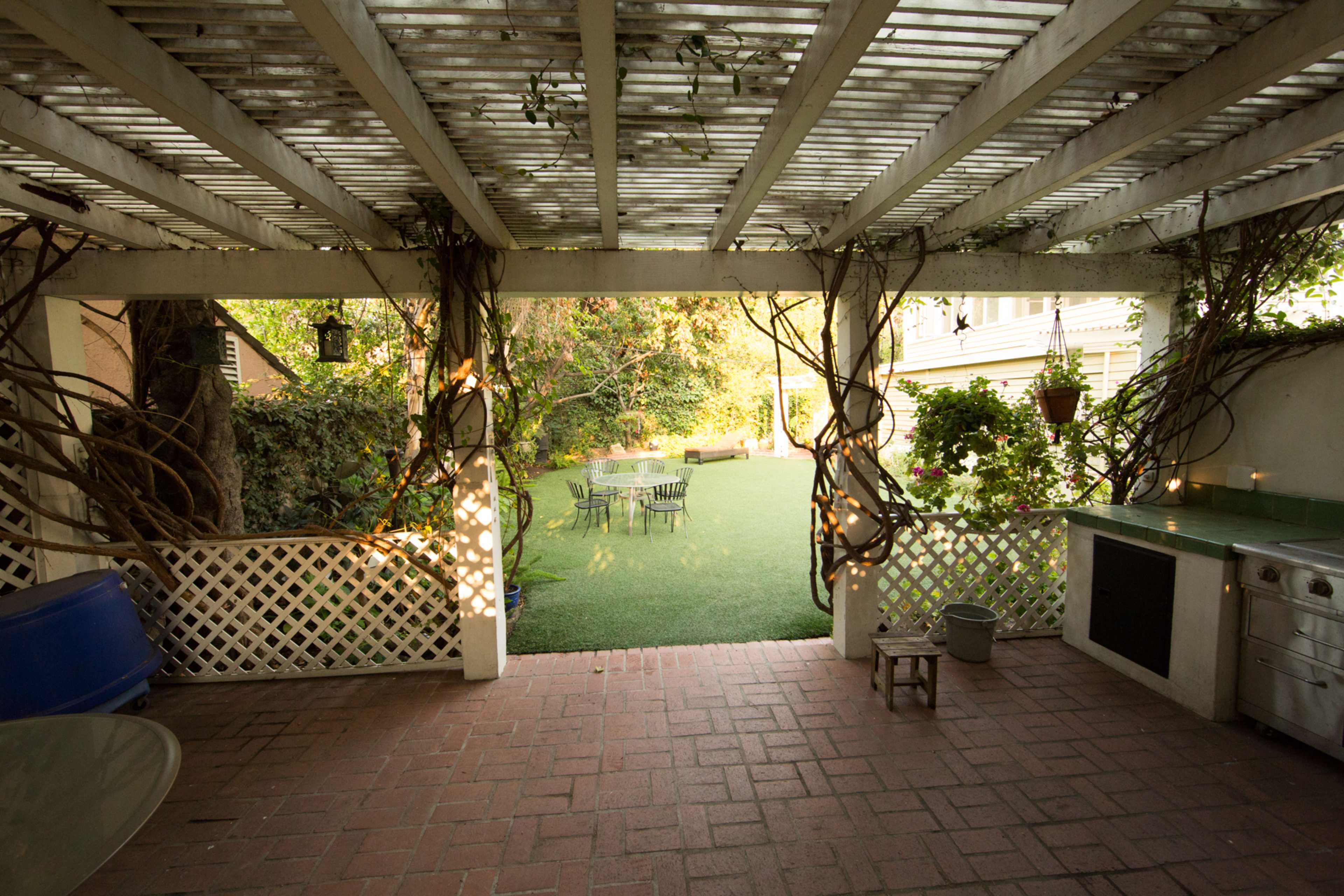 A covered outdoor patio with a brick floor, surrounded by greenery and a small dining area in an adjacent yard.