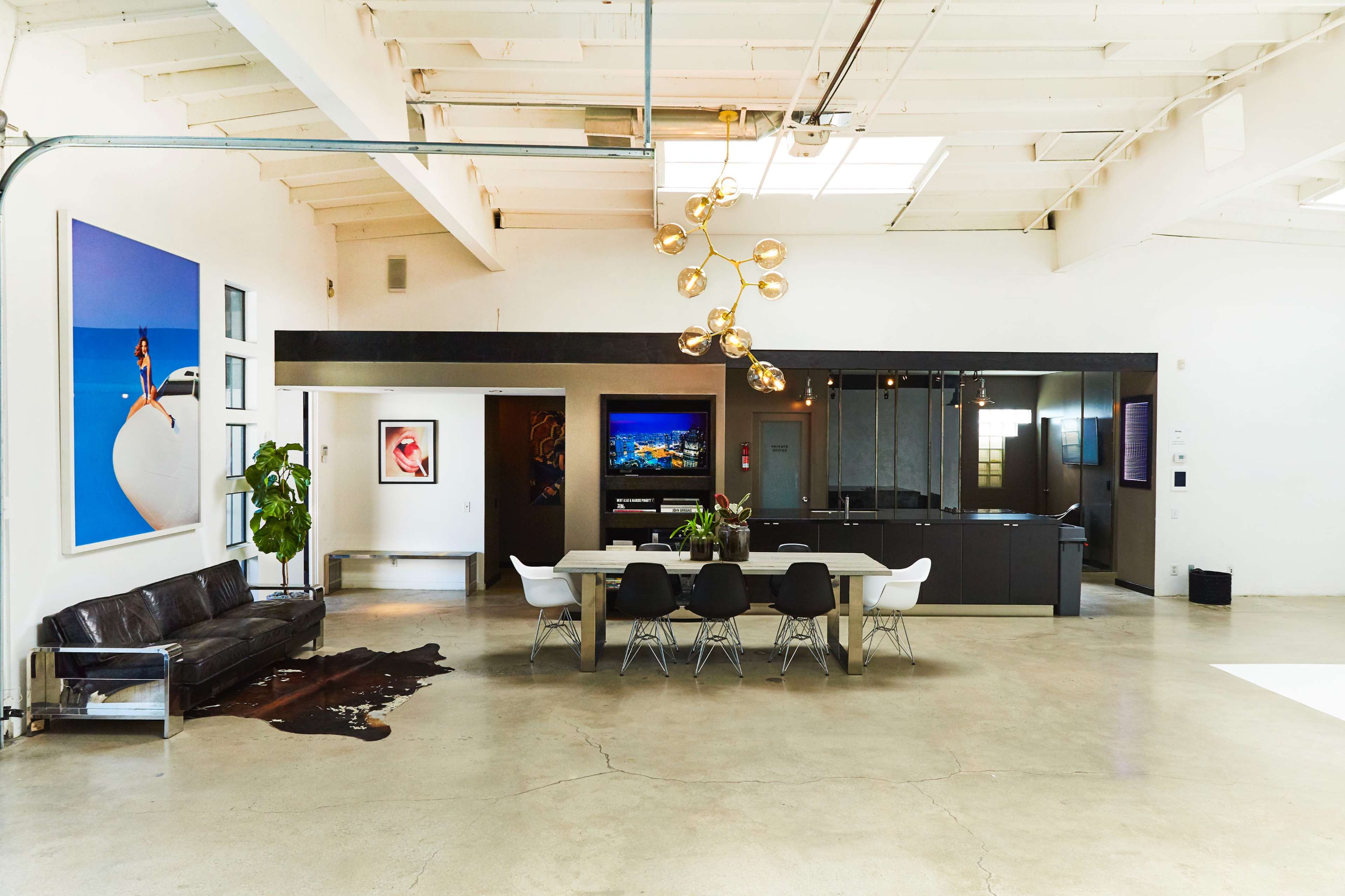 The image shows a modern, open-plan living space with a long dining table, a black leather sofa, large artwork on the walls, and a kitchen area in the background.