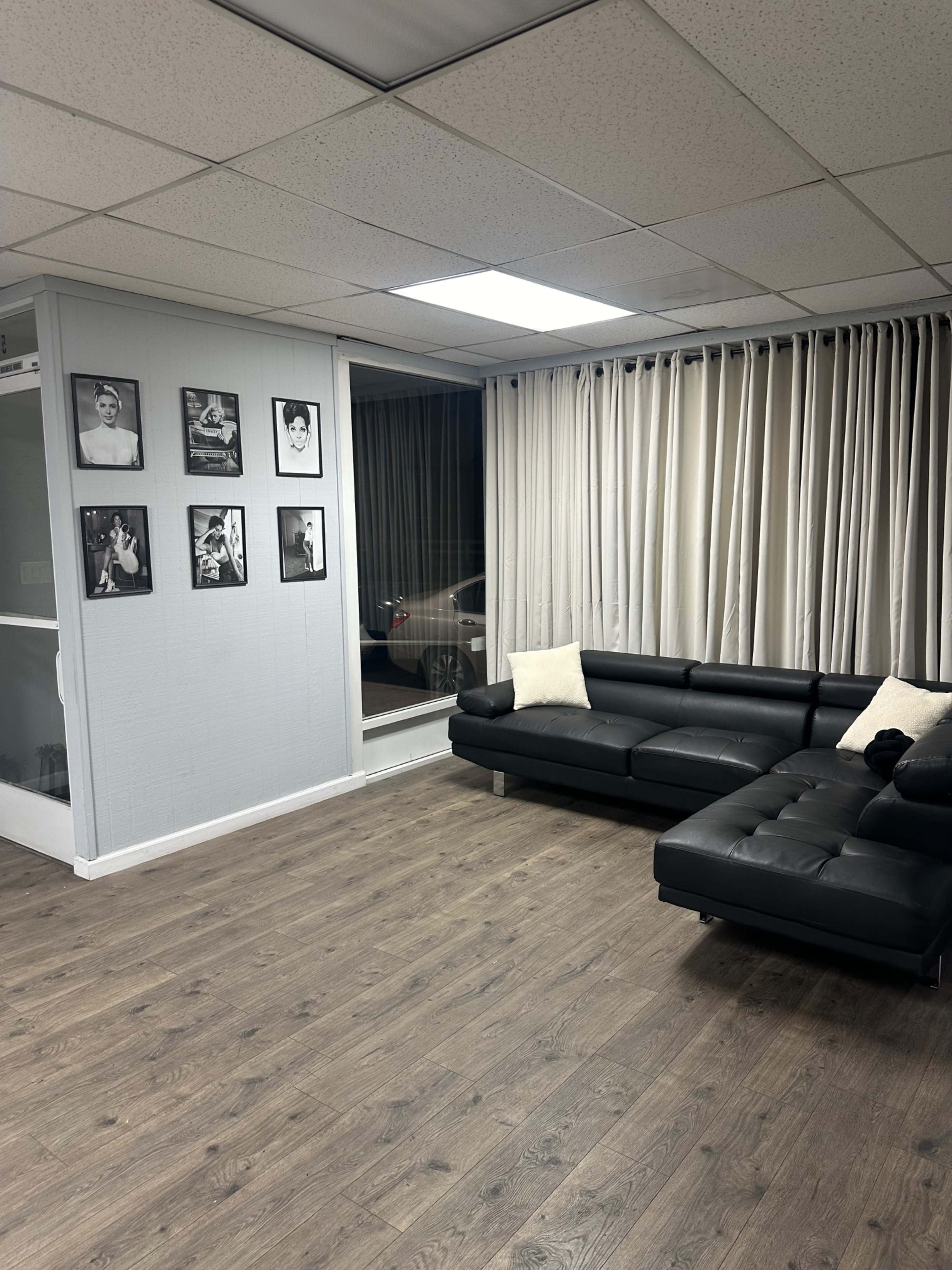 The image shows a well-lit, spacious room with dark wooden flooring, featuring a black leather sofa and several framed black-and-white photographs on the wall.