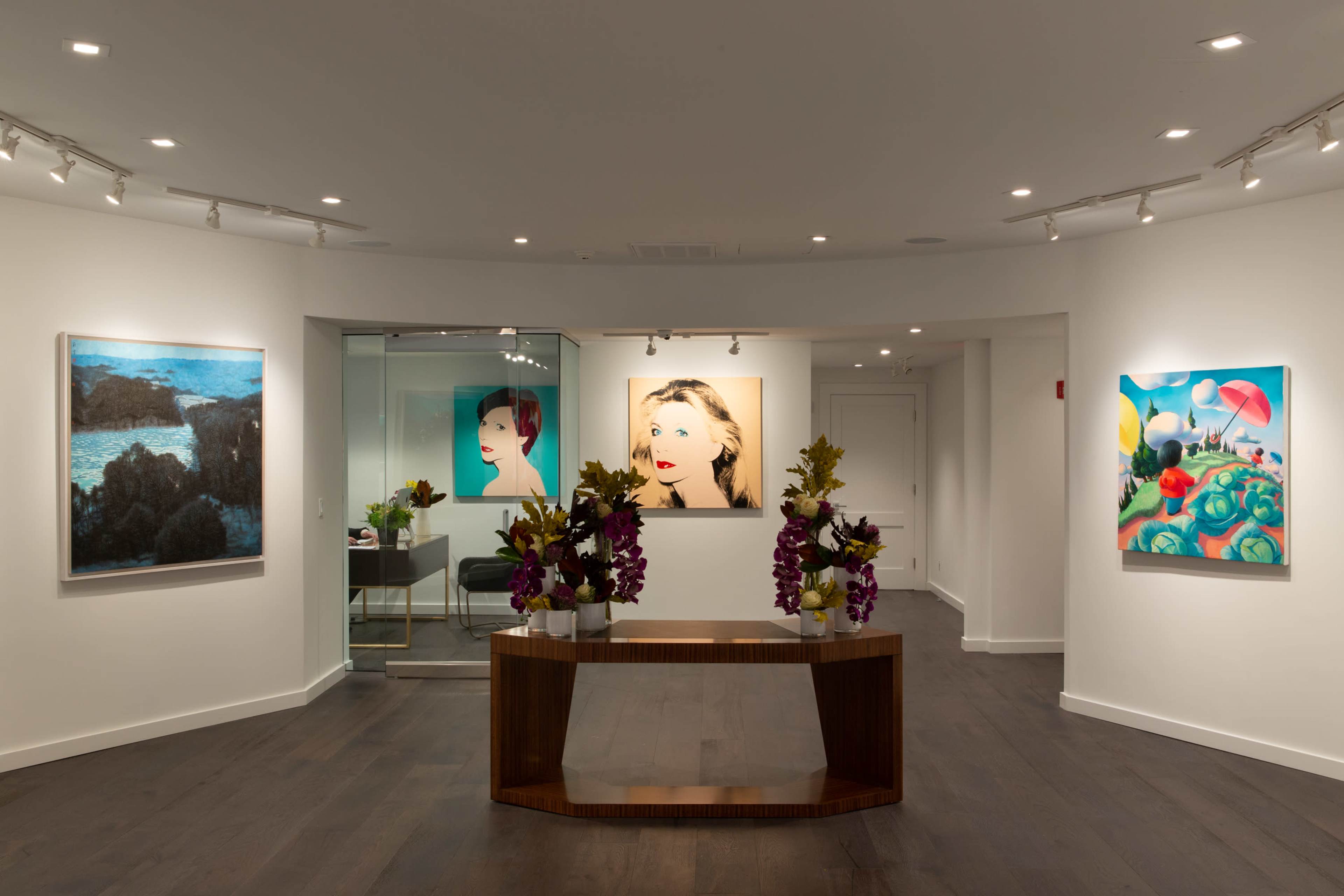 A modern art gallery featuring three paintings on the walls, a wooden table with floral arrangements in the center, and a mirrored area in the background.