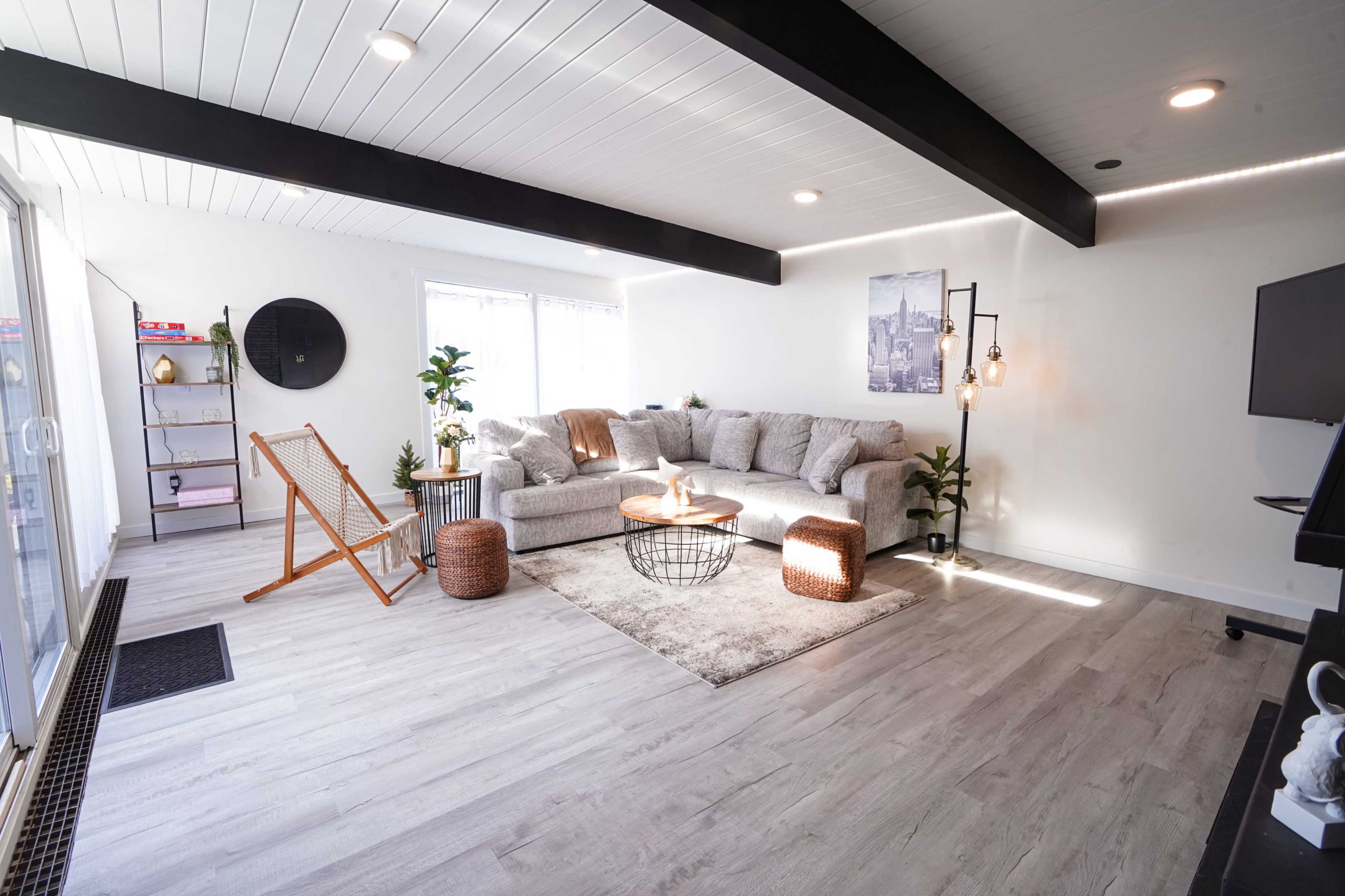 A light-filled living room with a large, gray sectional sofa, a coffee table, and two plants, featuring wood beam ceilings and modern decor.