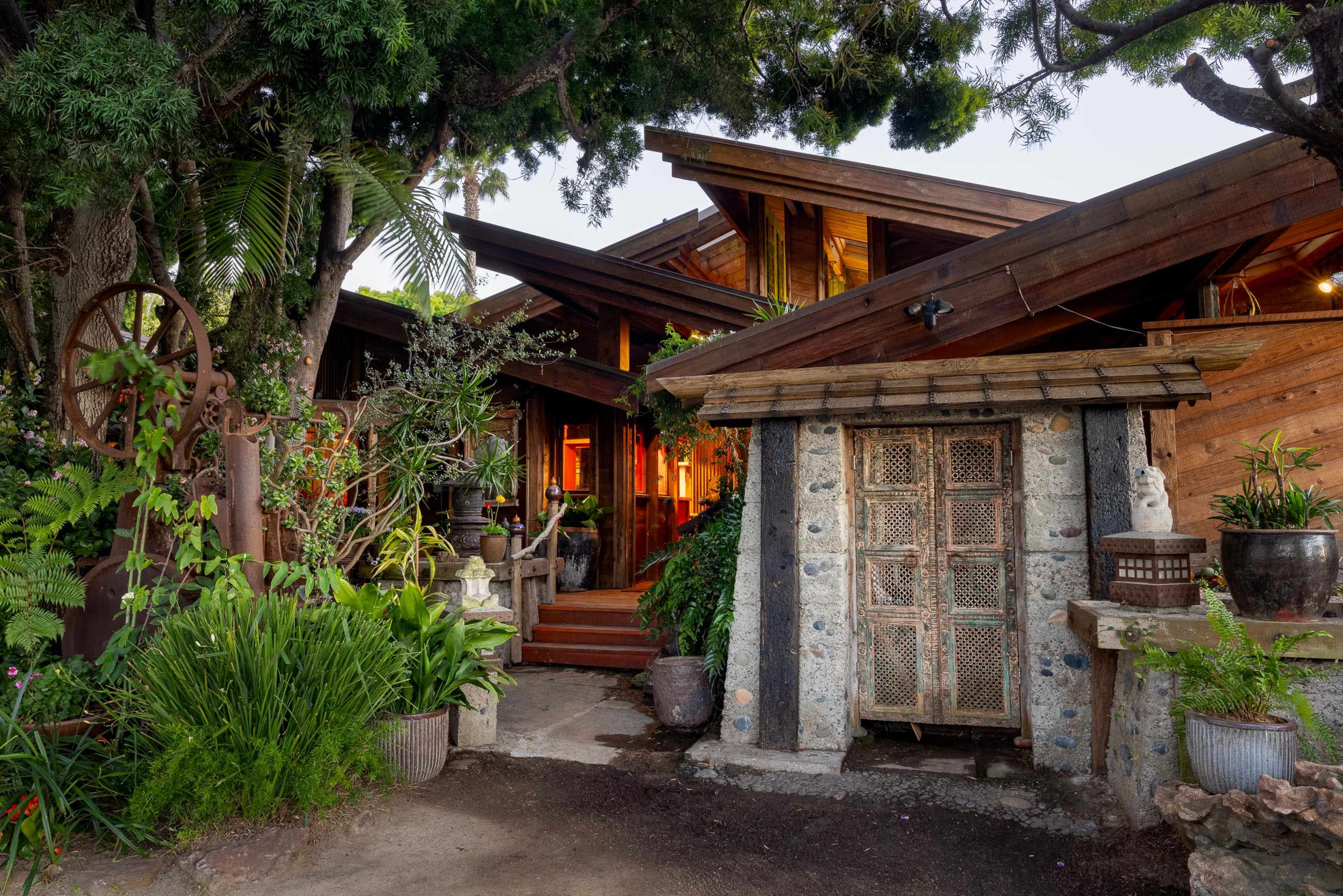 The image shows a rustic outdoor entrance to a wooden and stone building, surrounded by lush greenery and decorative elements.