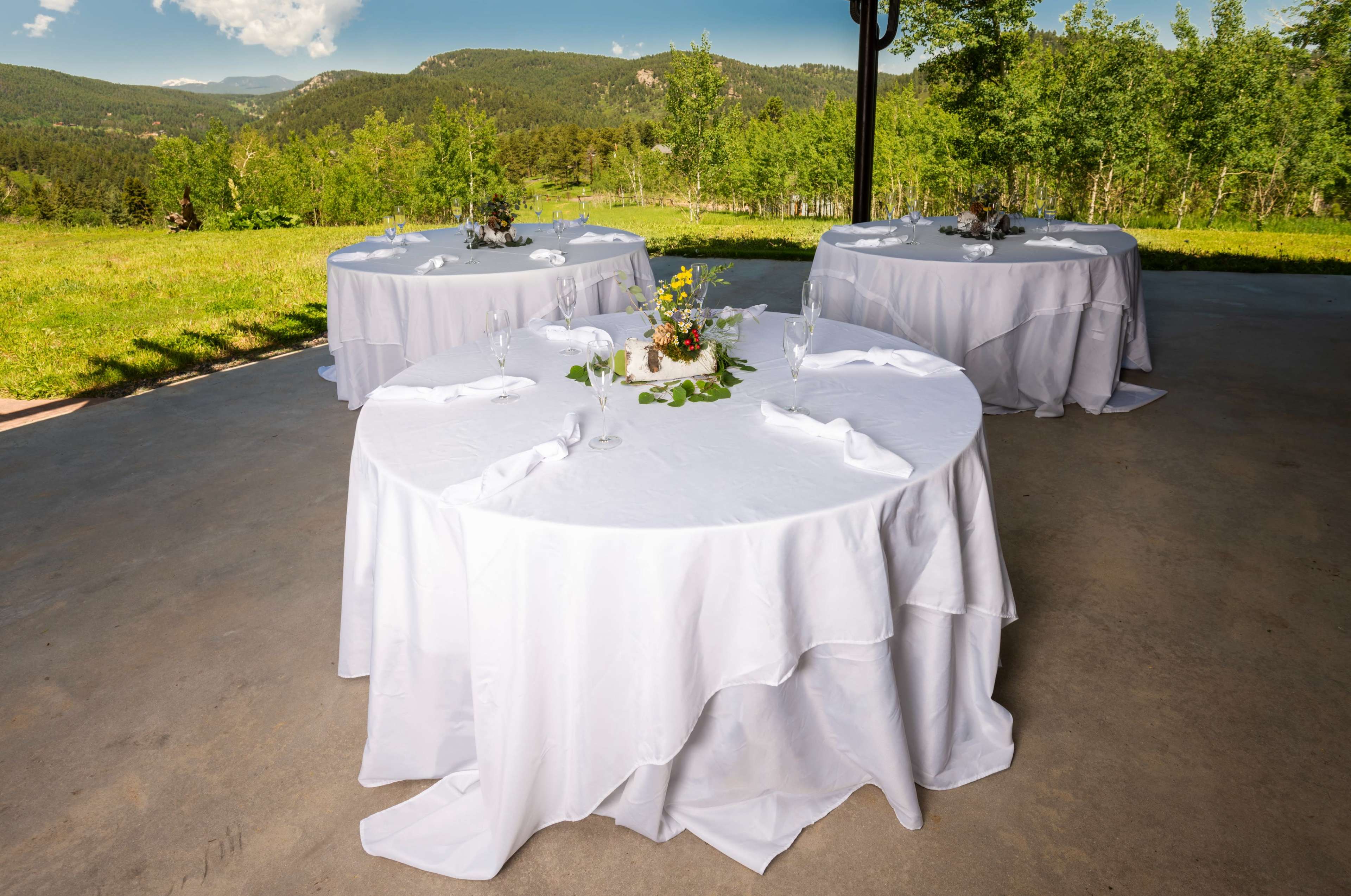 Three round tables covered with white tablecloths are arranged outdoors on a concrete surface, surrounded by lush greenery and mountains in the background.