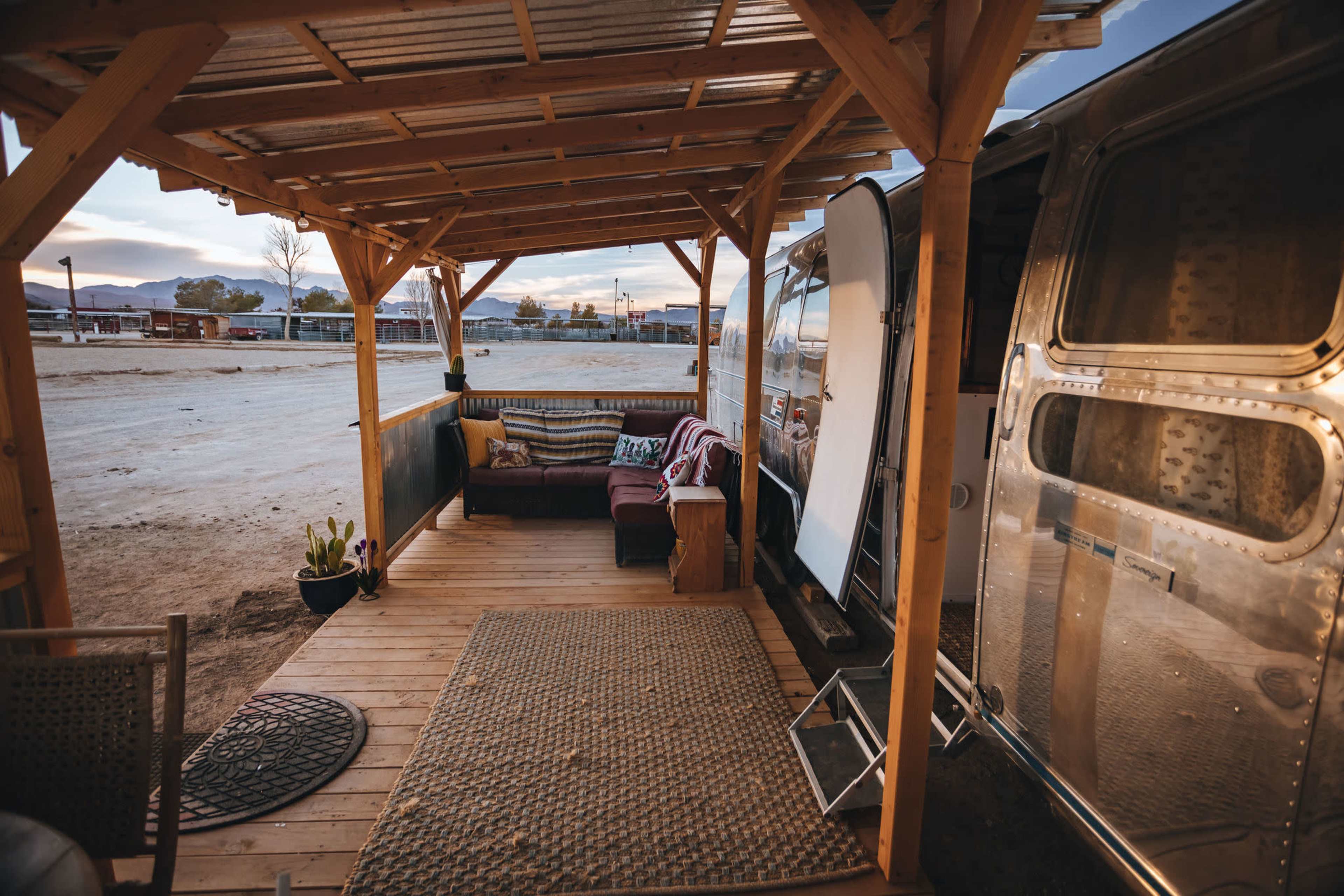 A wooden porch with seating and a rug beside an Airstream trailer in an open, rural area.