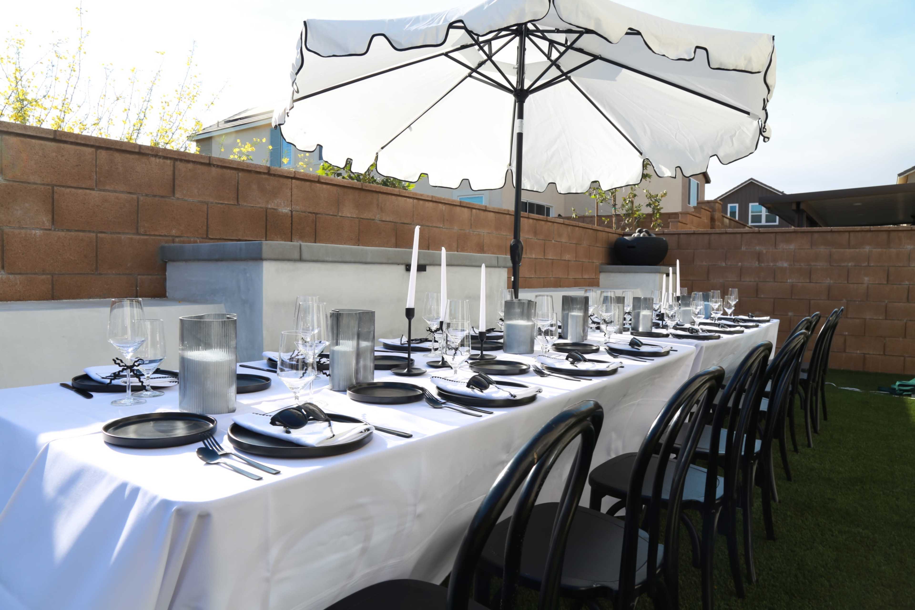 A long dining table is set for a meal, featuring black plates, glassware, and candles, under a white umbrella in a garden space with a brick wall backdrop.