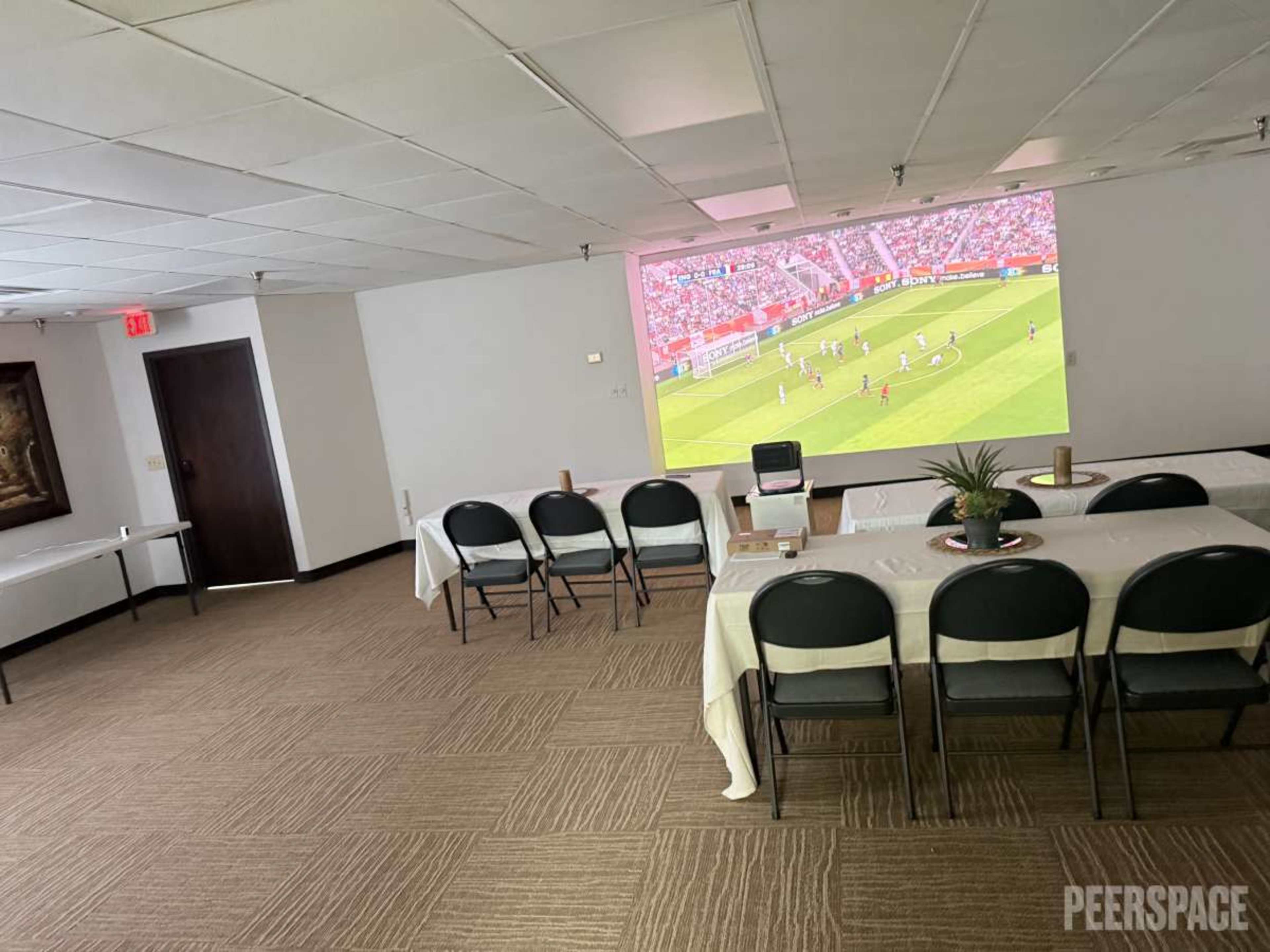 A large room features a projector displaying a soccer match, with several tables set up and chairs arranged for an audience.