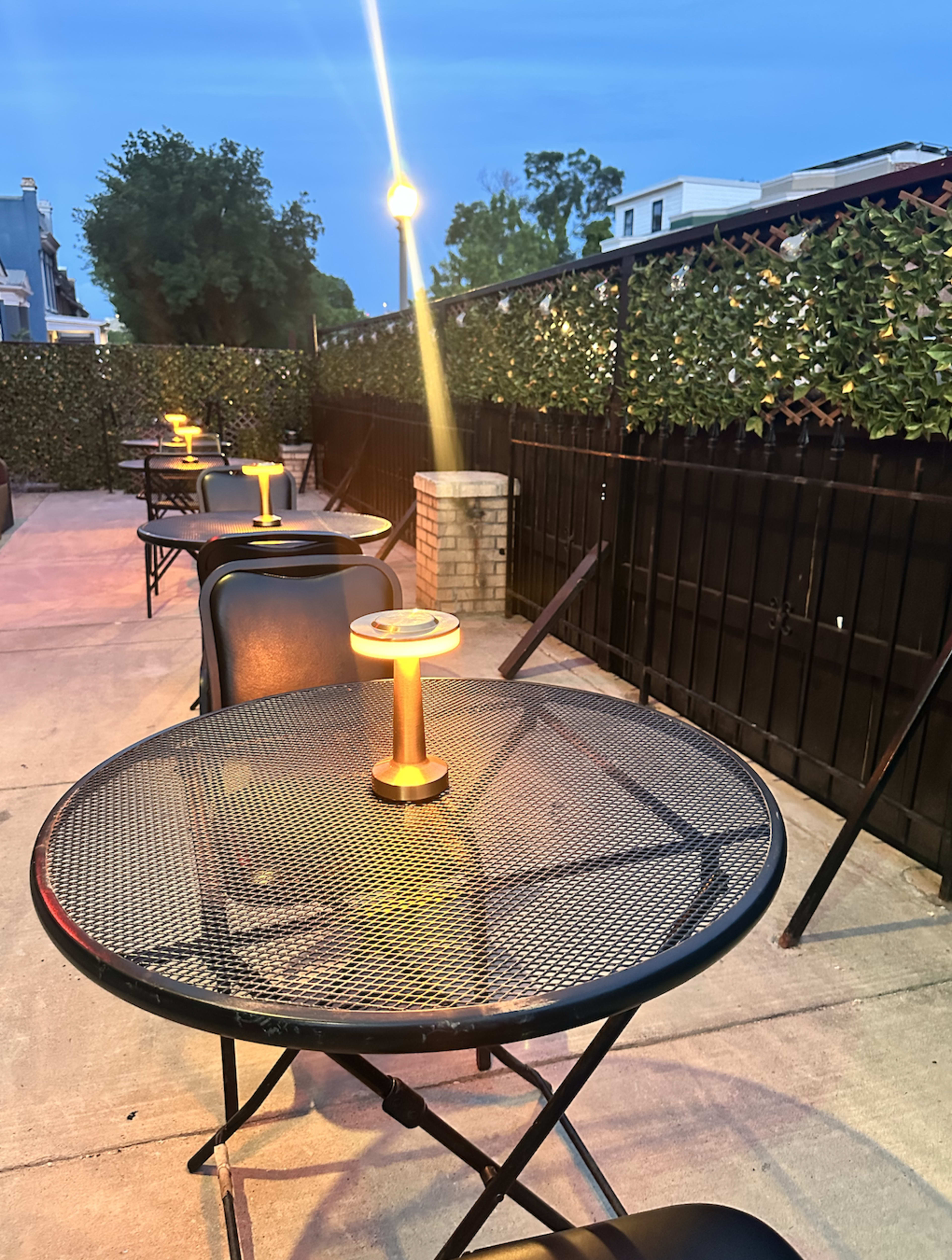 A series of empty metal tables with gold candle holders line a concrete patio, surrounded by a tall black fence and greenery under a dusky blue sky.