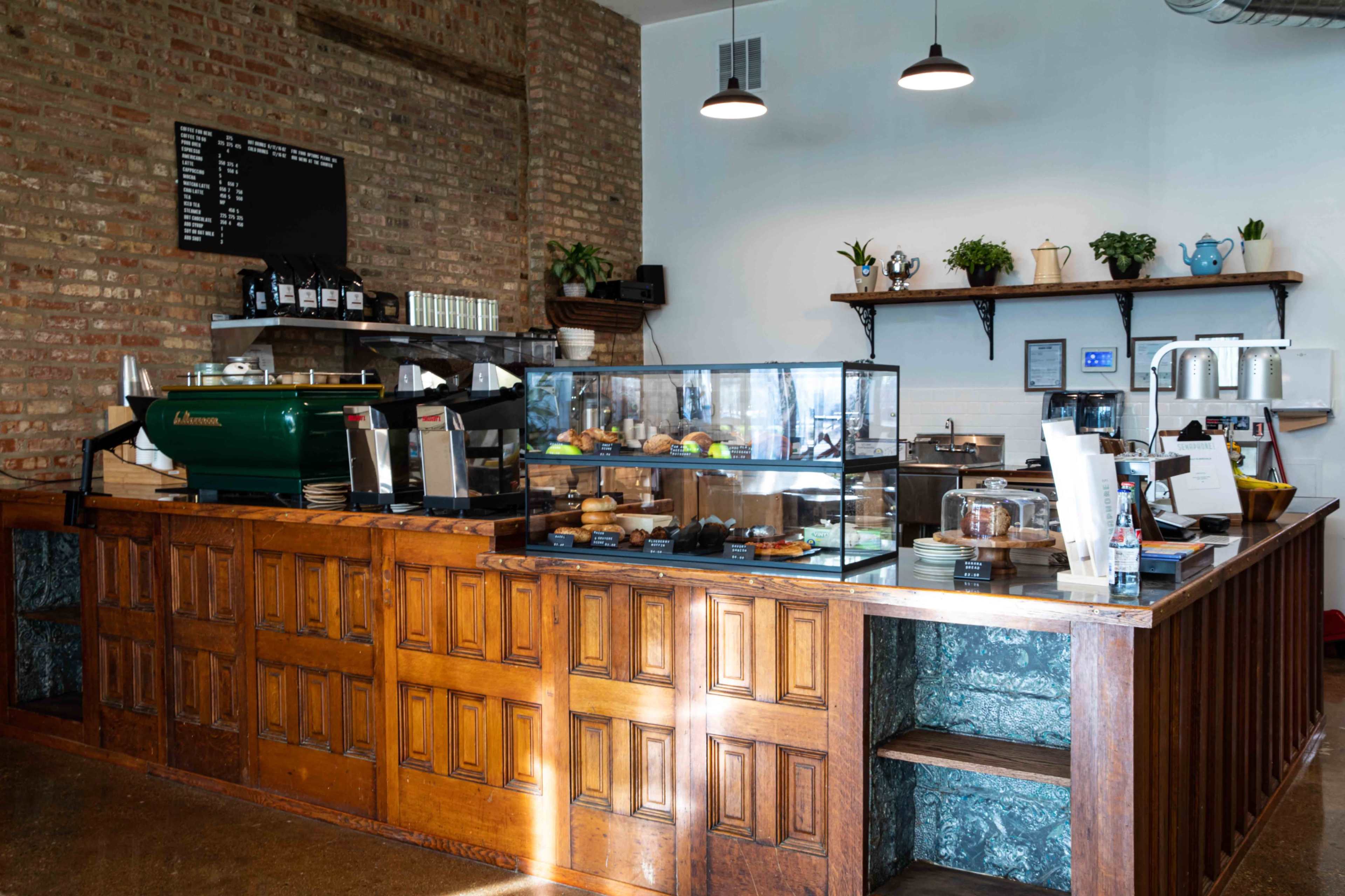 The image shows a coffee shop counter featuring a glass display case with baked goods, a coffee machine, and a wooden service area with a menu board on the wall.