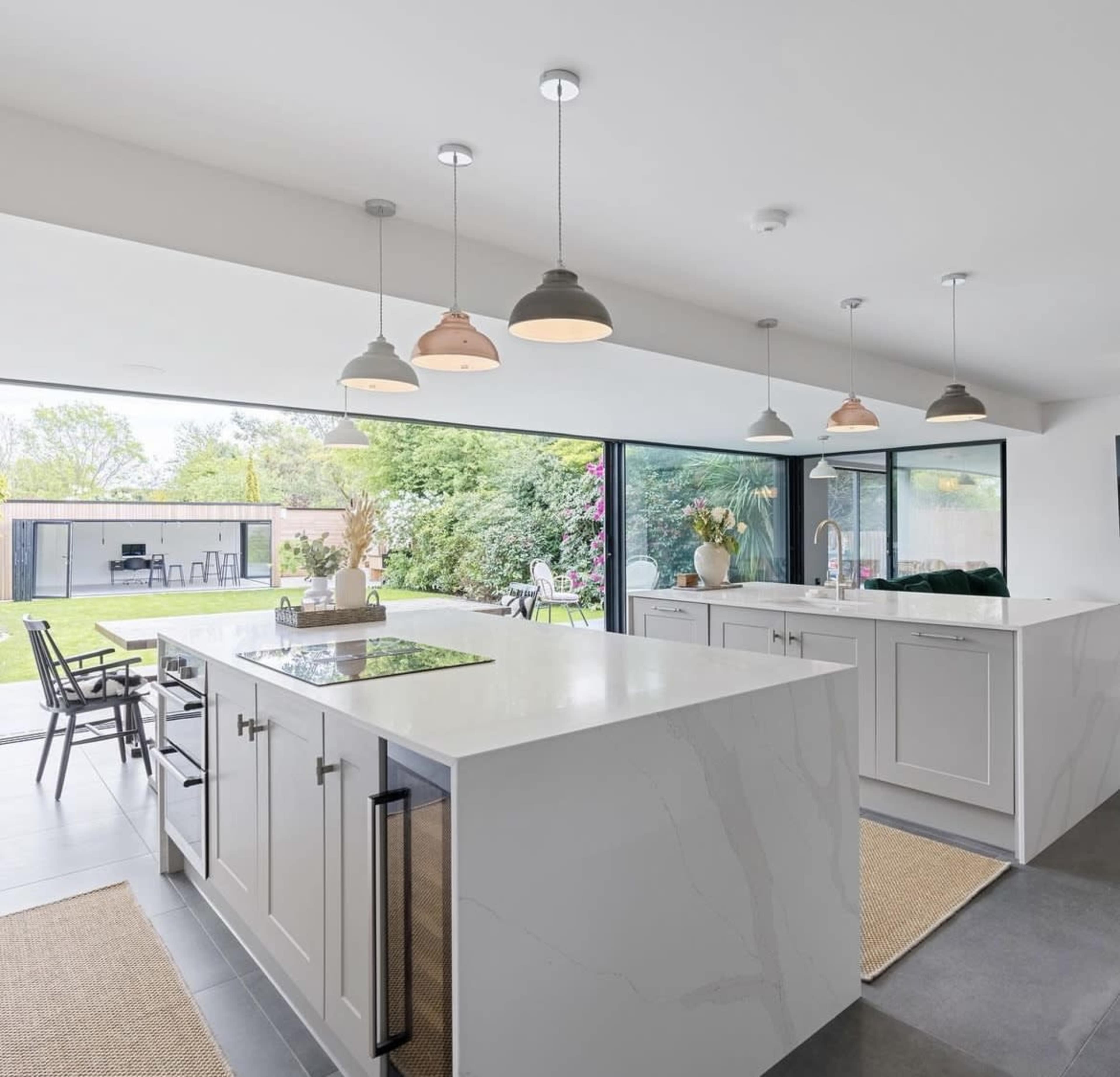 A modern kitchen features a large island with a cooktop, cabinets, and pendant lights, opening to a landscaped backyard through expansive glass doors.
