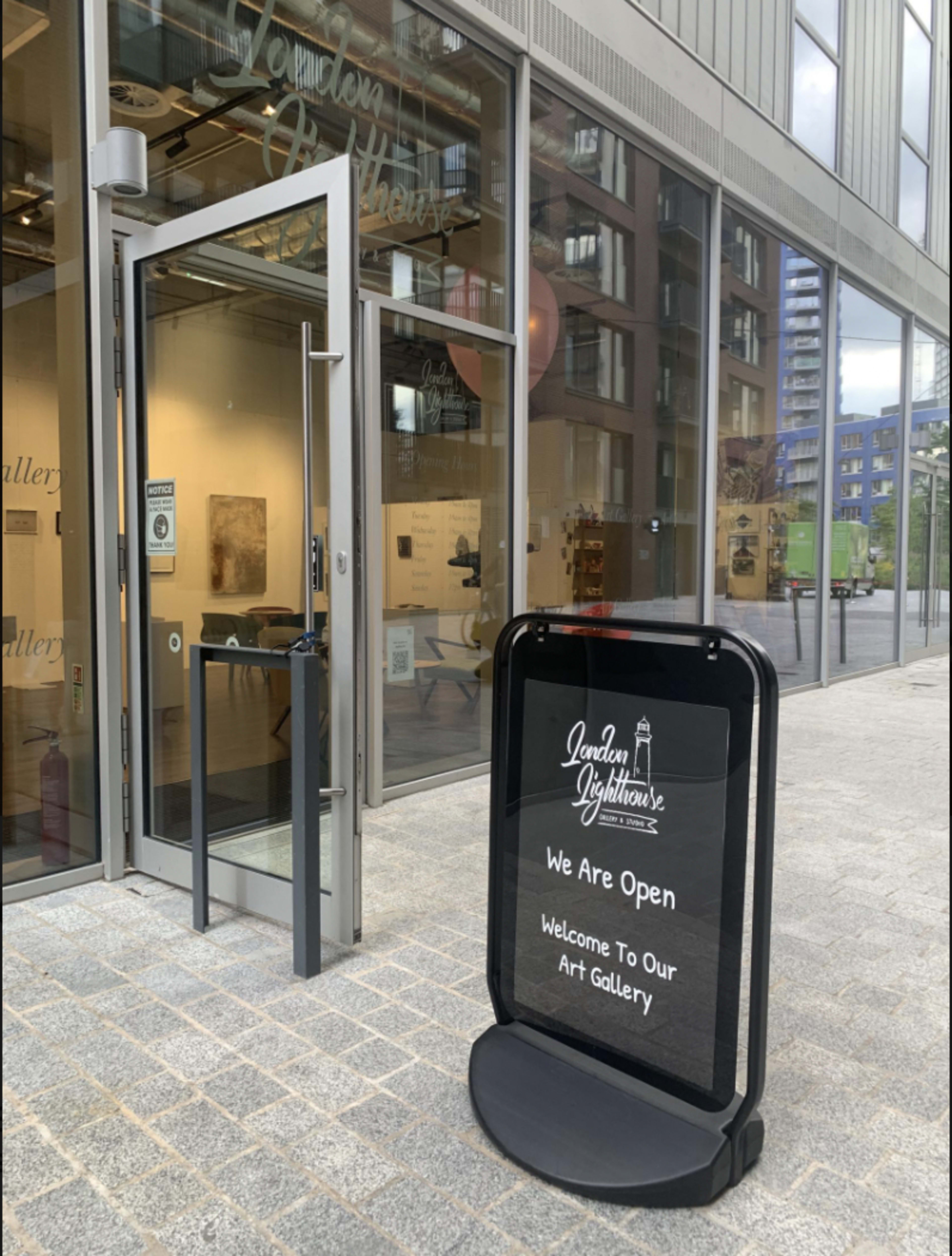 A black sandwich board sign at the entrance of an art gallery displays a message welcoming visitors and indicating that the gallery is open.