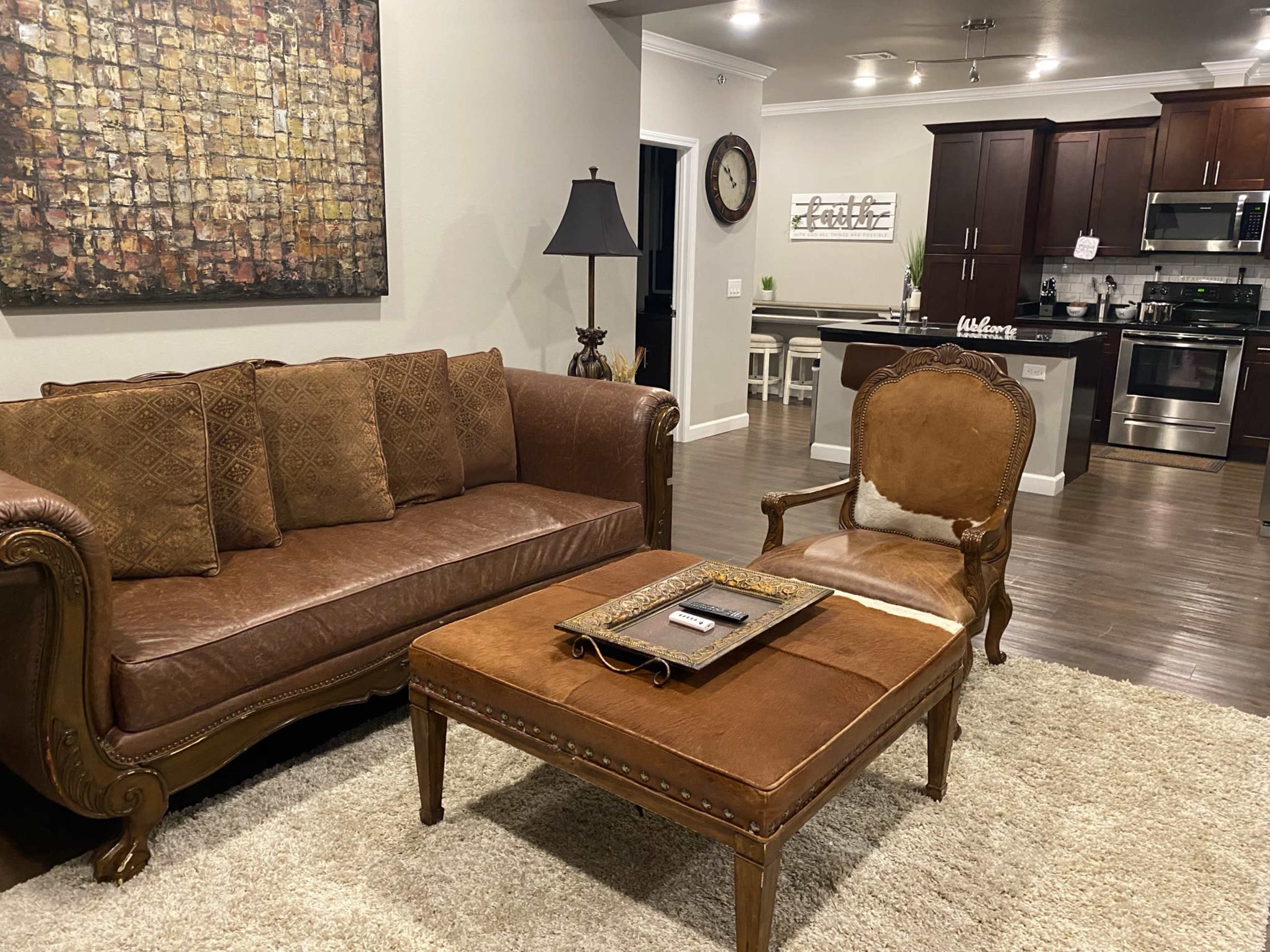 A living room featuring a brown leather sofa, an ornate chair, a wooden coffee table, and a kitchen area in the background with dark cabinets and stainless steel appliances.