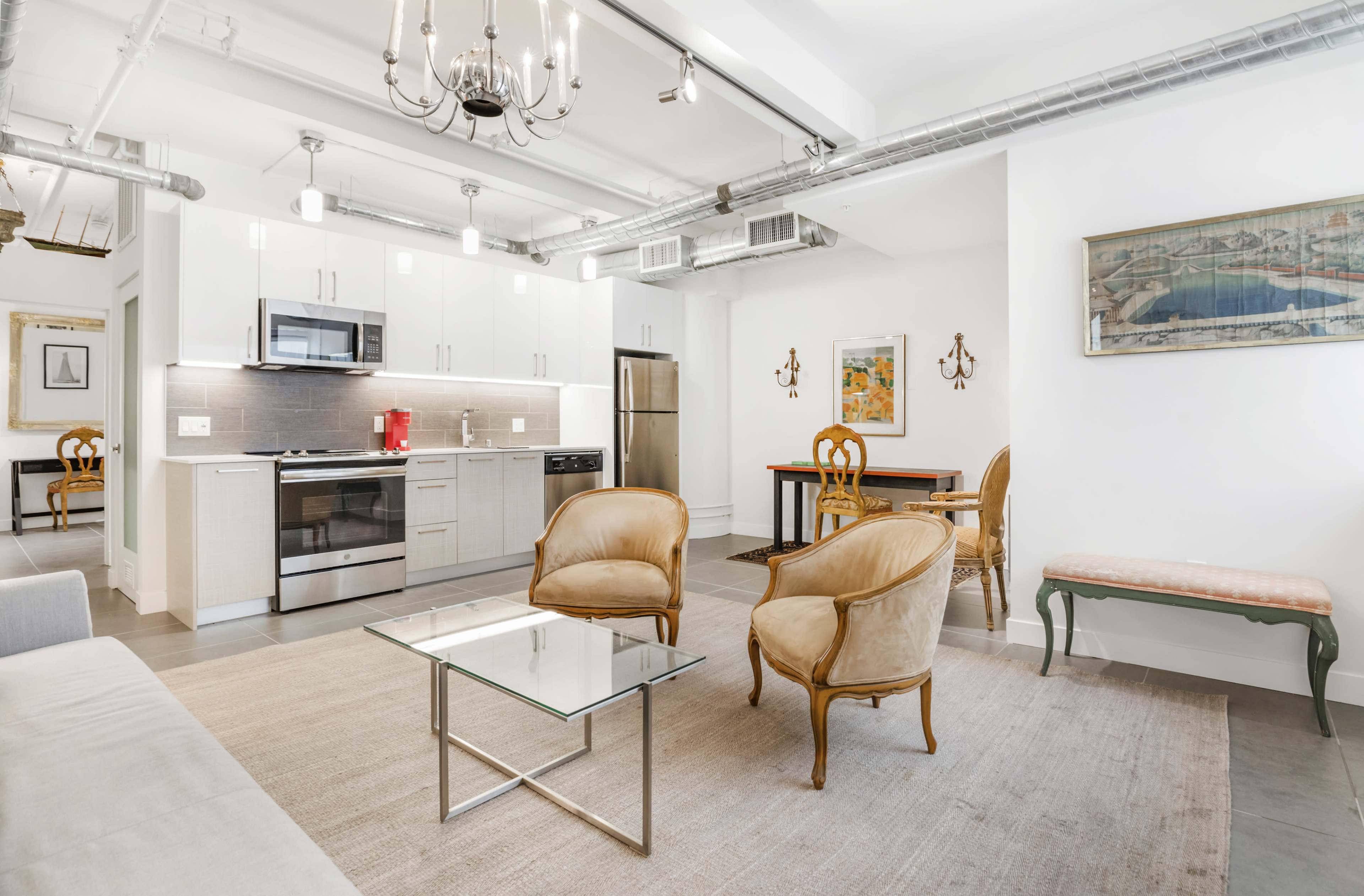 The image shows a modern, open-concept kitchen and living area with a dining table, two armchairs, and a decorative chandelier.