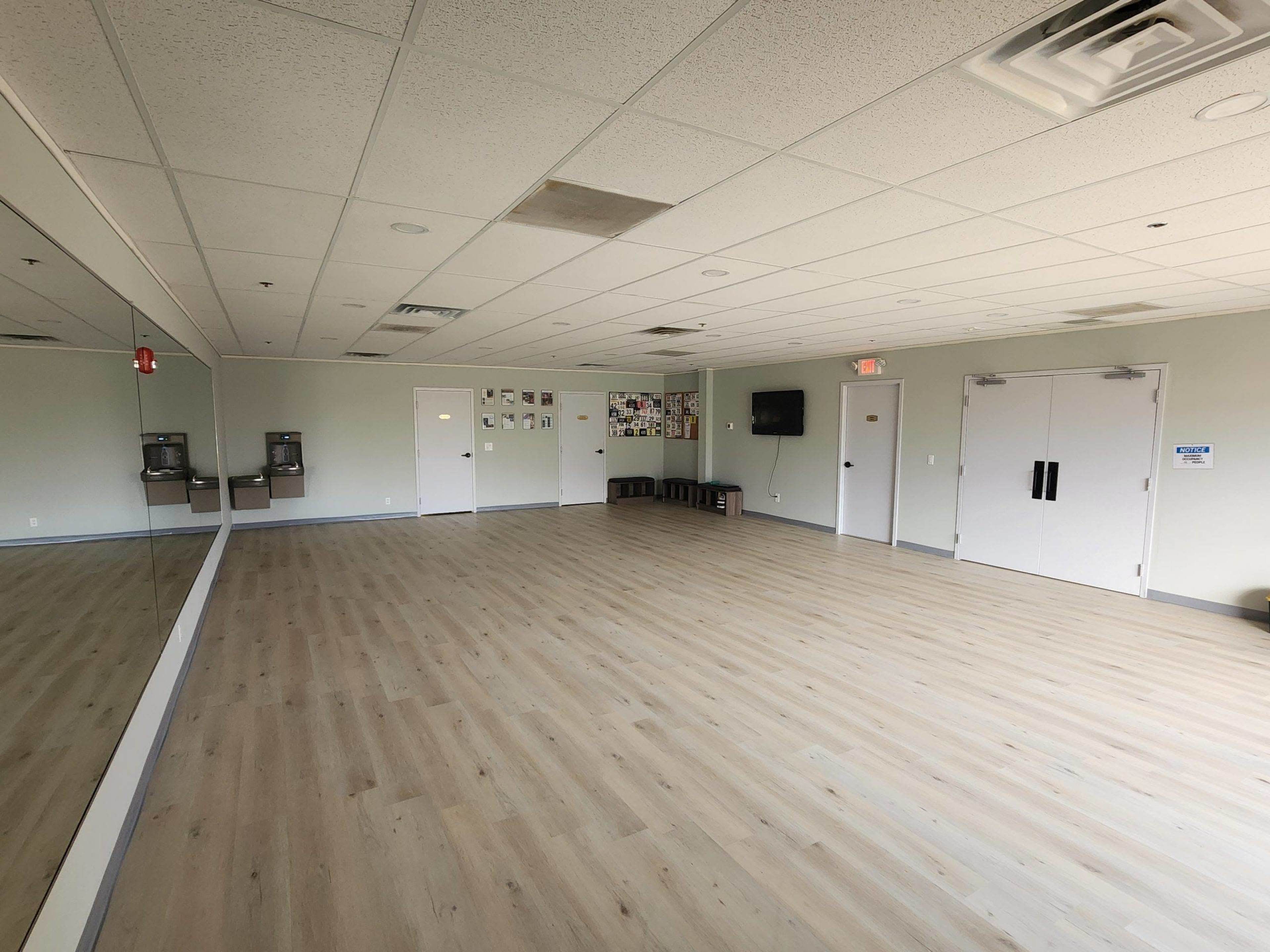 The image shows a spacious, empty room with wooden flooring, large mirrors on one wall, and a small kitchenette area with a door and a television mounted on the opposite wall.