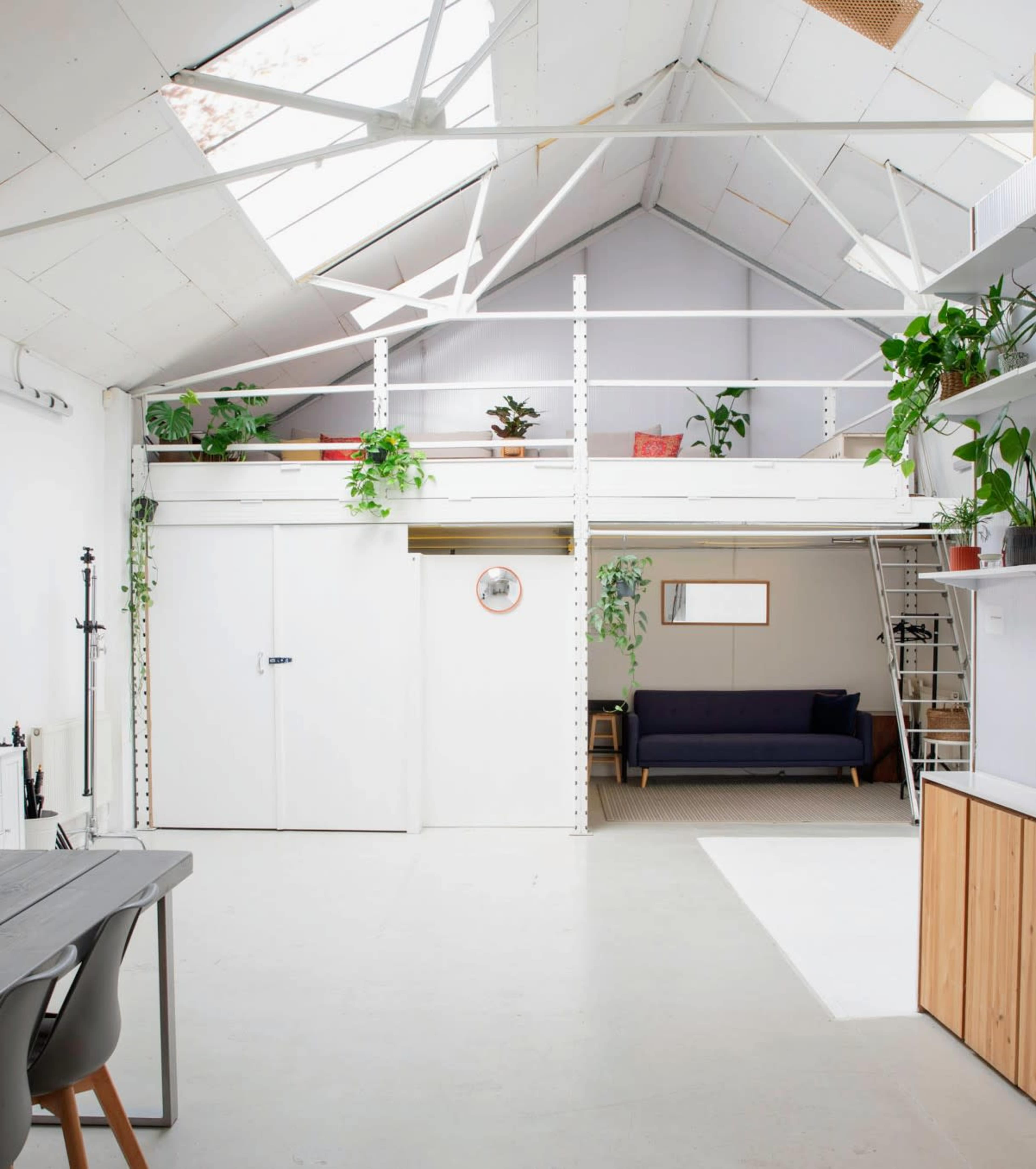 The image shows a modern, bright interior of a loft space with a mezzanine, featuring plants, a sofa, and a dining area.