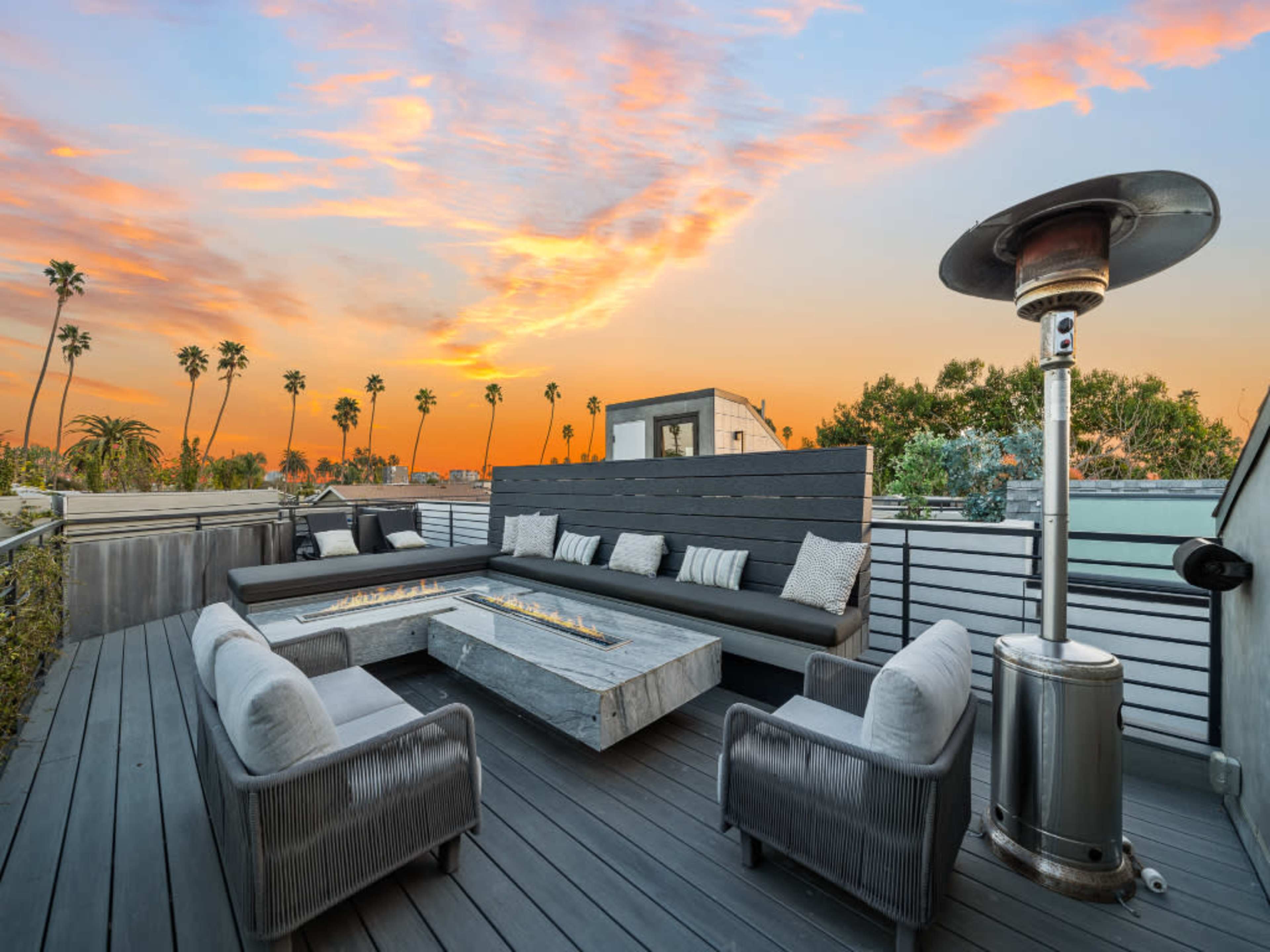 The image shows a rooftop terrace with a modern seating area, fire pit, and a view of palm trees against a colorful sunset sky.
