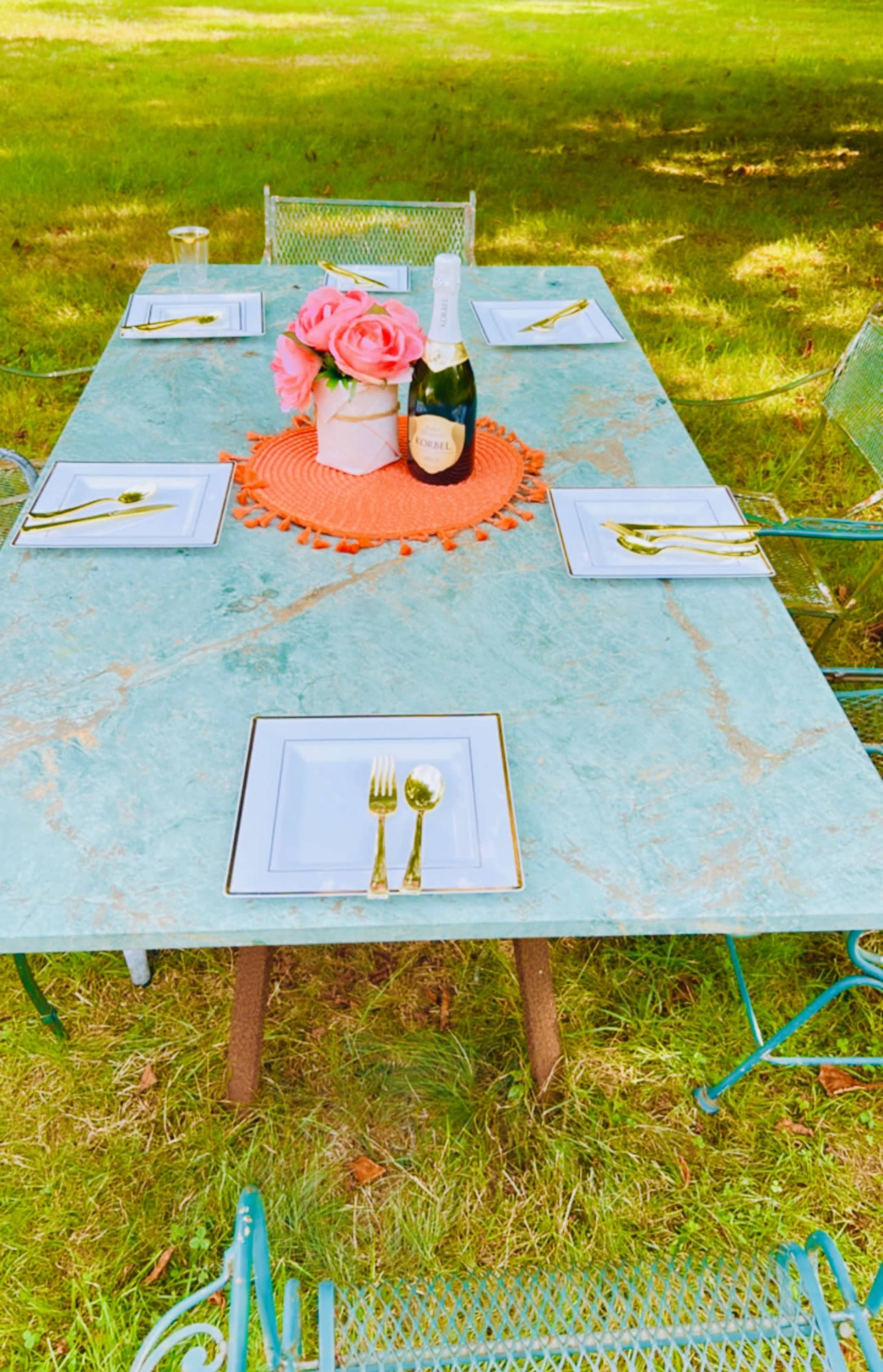 A marble table is set with gold utensils and white plates, adorned with a floral centerpiece and a bottle of champagne, surrounded by chairs on a grassy area.