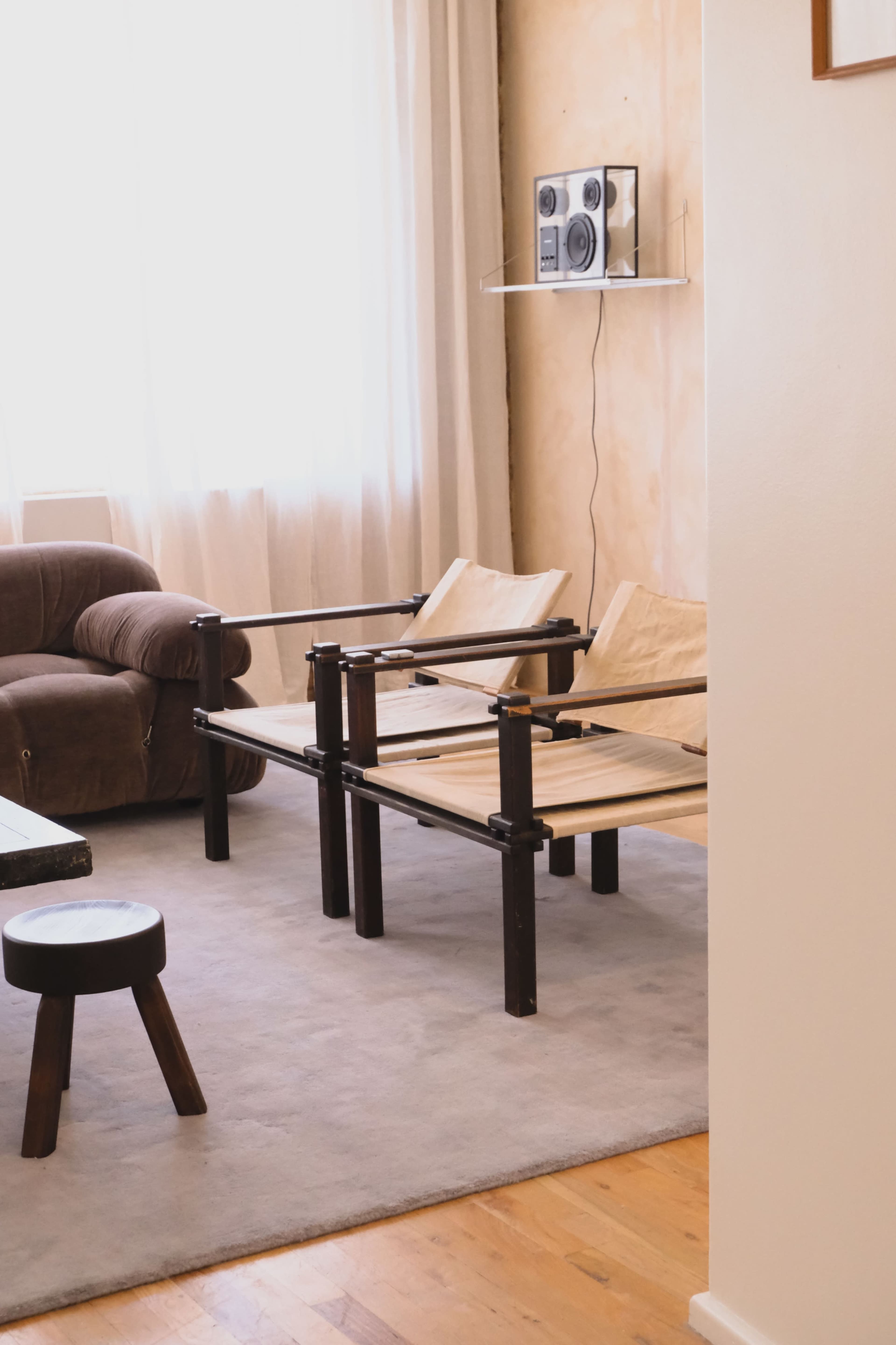 The image shows a modern living room with two wooden chairs, a brown sofa, a side table, and a small audio system mounted on the wall.