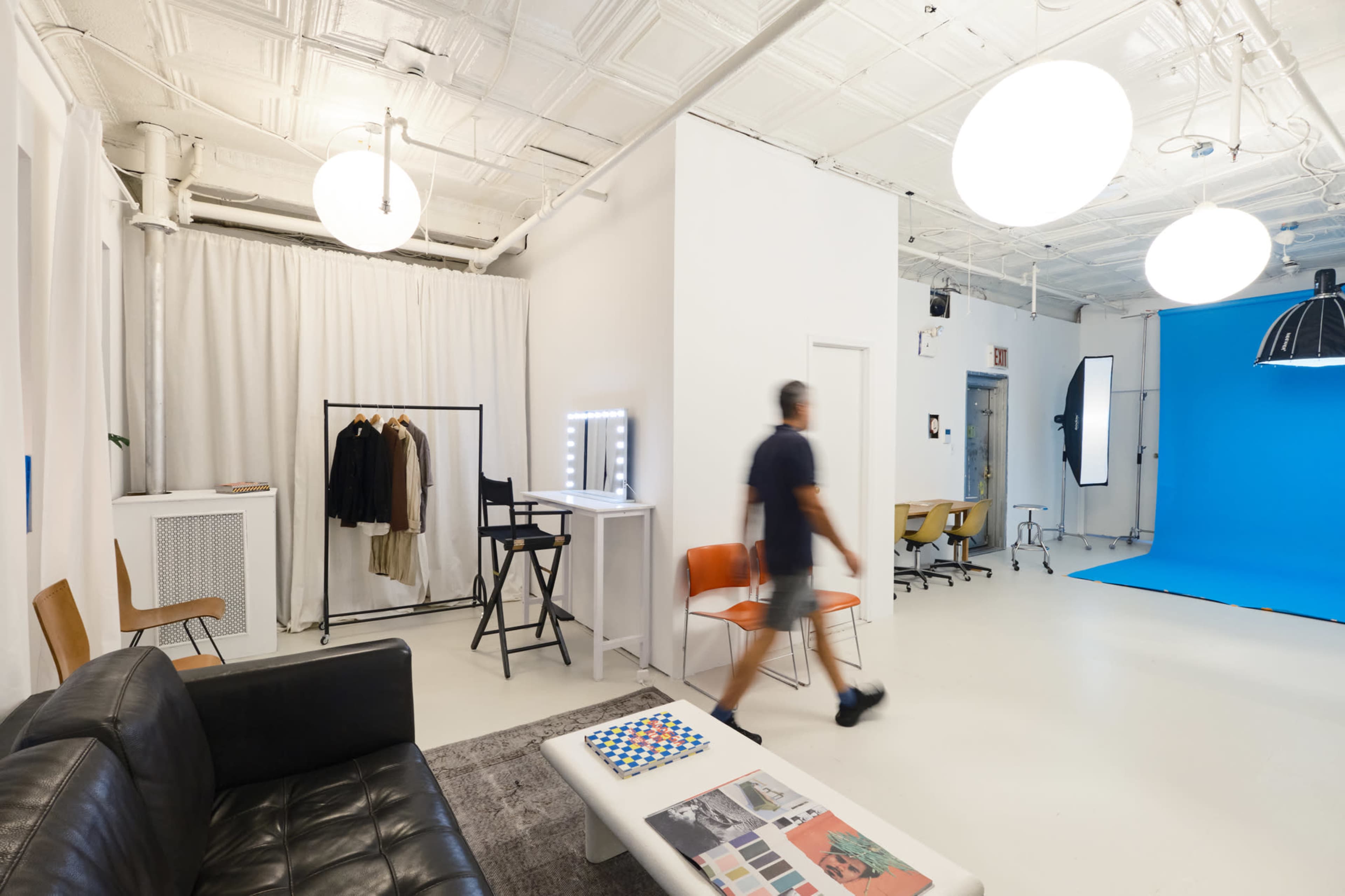 The image depicts a brightly lit studio space with a blue backdrop, various chairs, and clothing on display, featuring a person walking through the area.