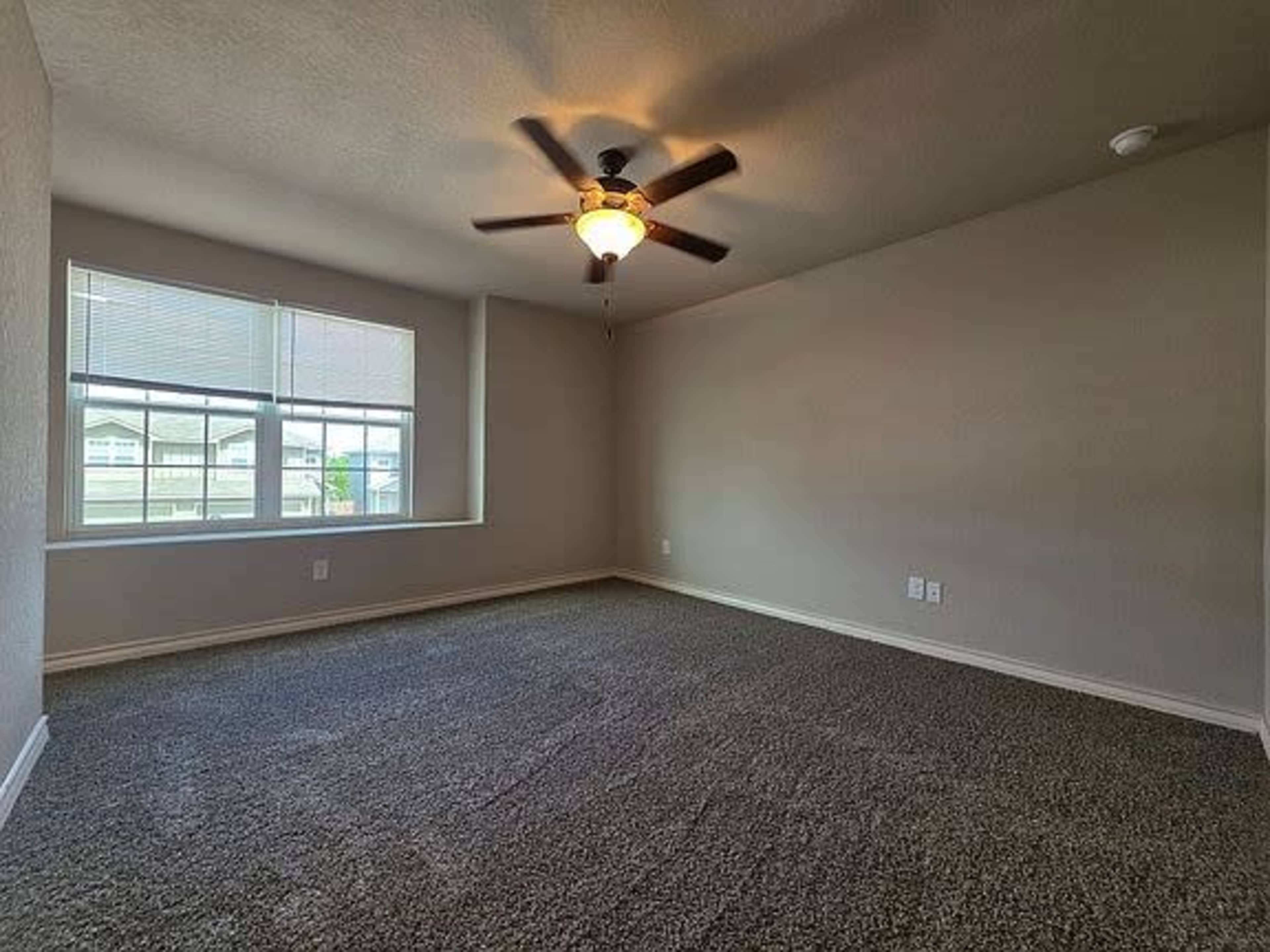 The image shows a spacious, empty room with gray carpet, a ceiling fan, and a large window allowing natural light to enter.