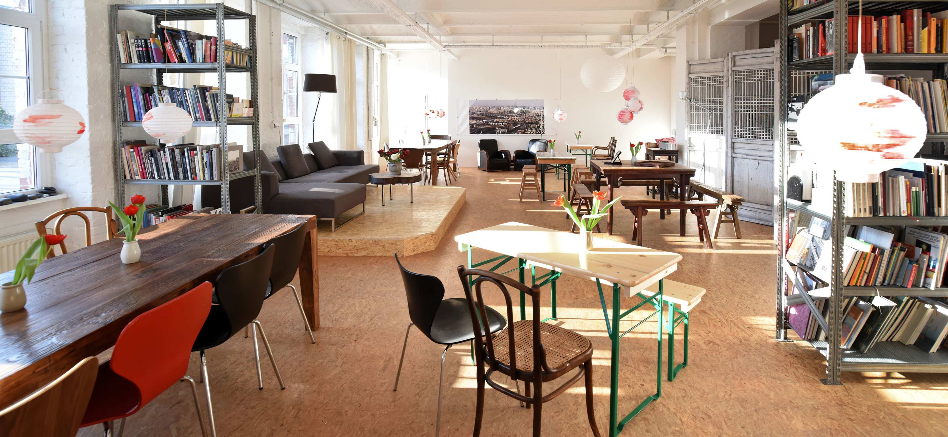 A bright, spacious room with wooden tables, chairs, and bookshelves, featuring a mix of seating areas and decorative elements like plants and hanging lights.