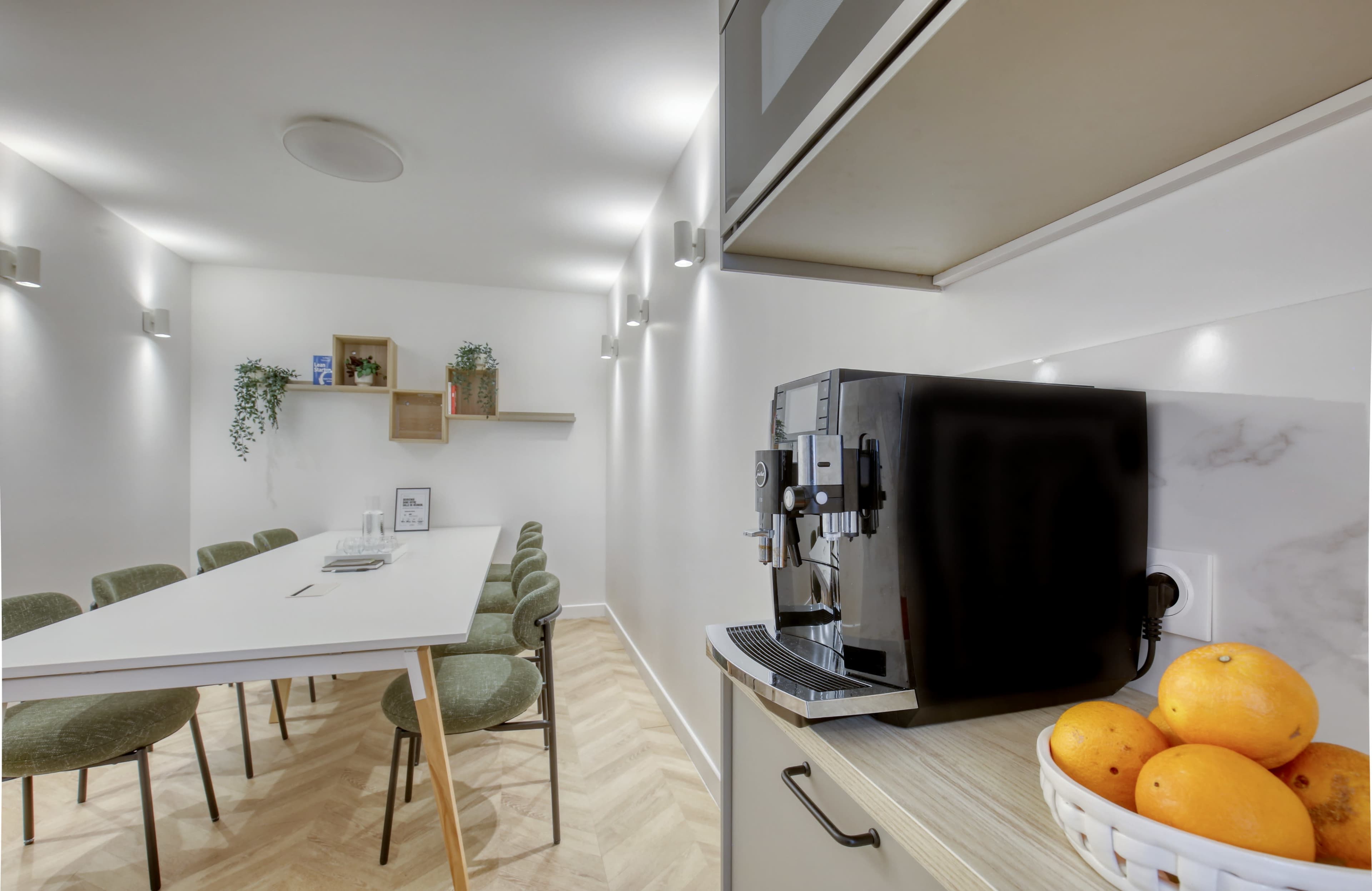 The image shows a modern kitchen with a coffee machine on the counter, a long table with chairs, and a bowl of oranges in a well-lit environment.