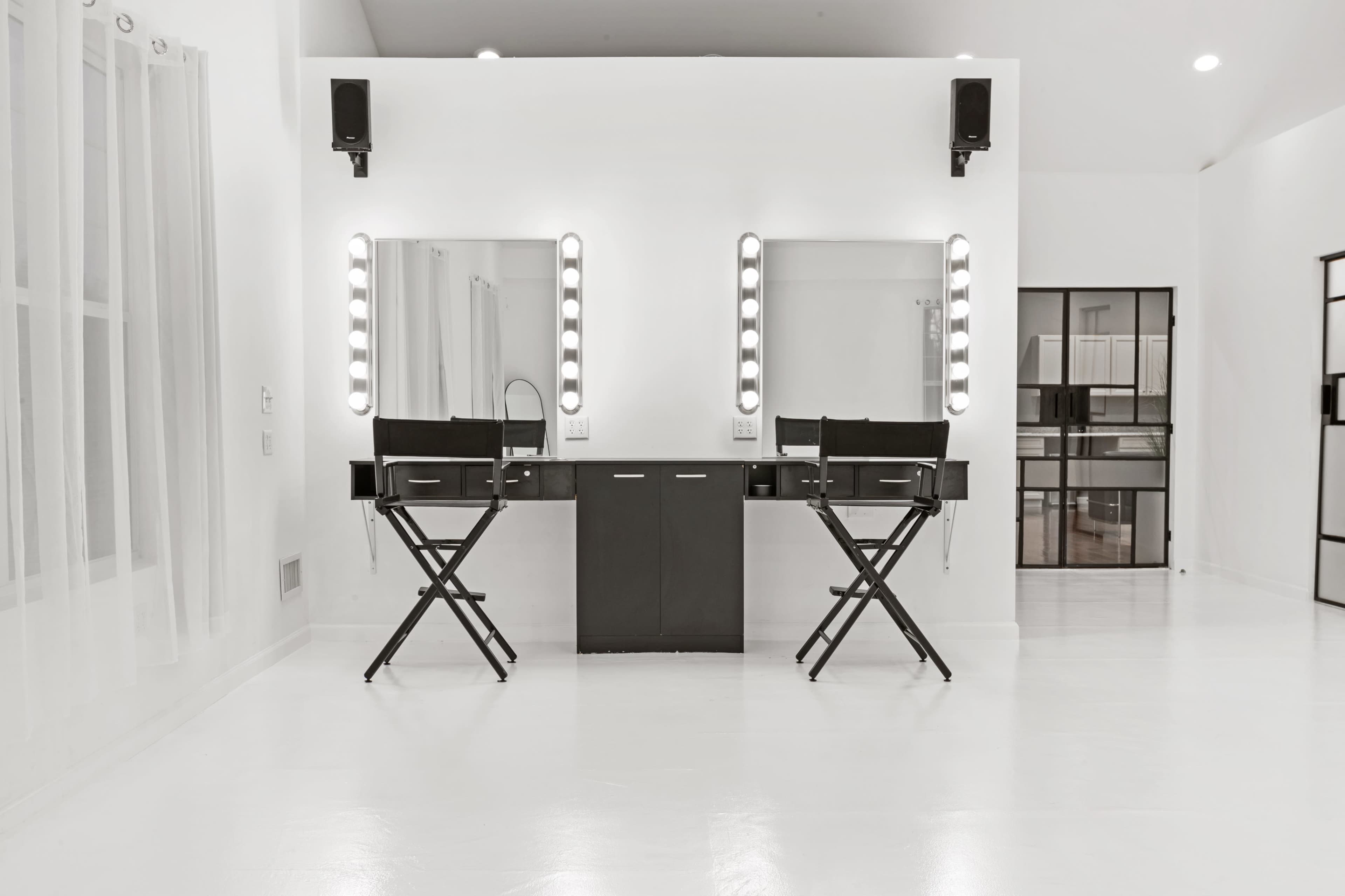 A minimalist beauty studio with two black makeup mirrors illuminated by light bulbs, flanked by matching director's chairs and a sleek black cabinet.