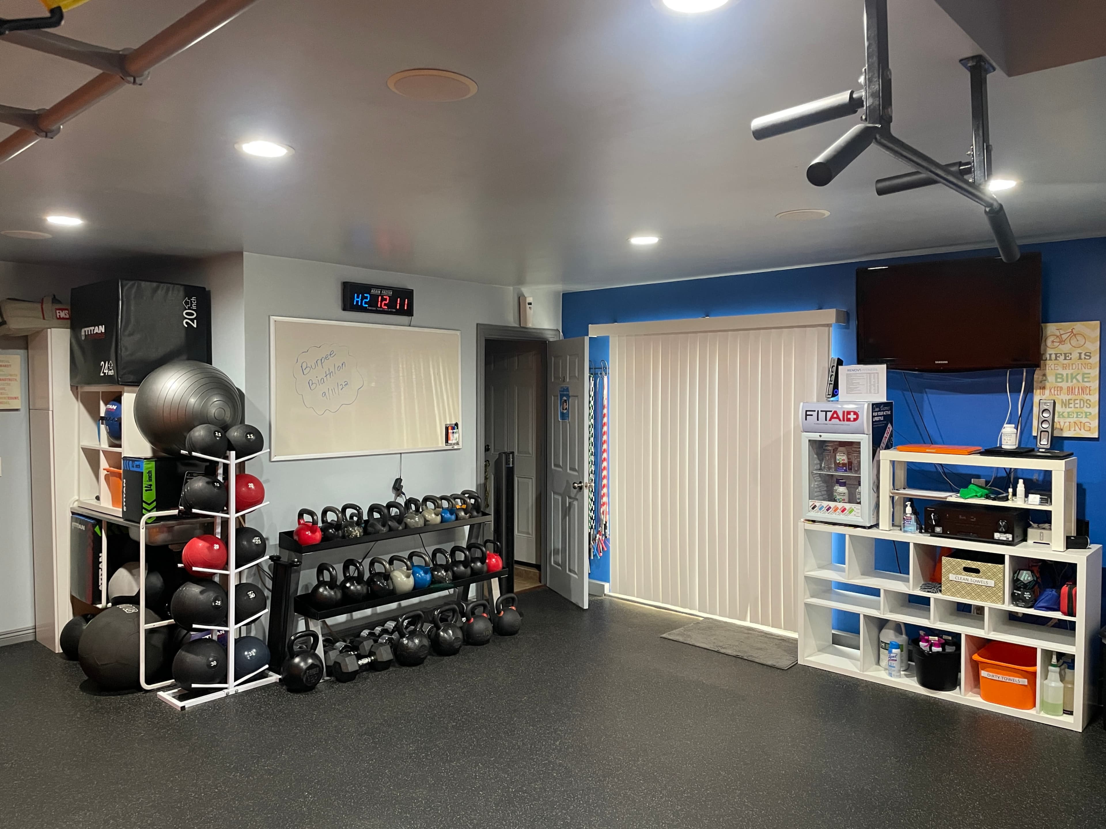A neatly organized home gym with a rack of kettlebells, exercise balls, and a wall-mounted television in a well-lit room.