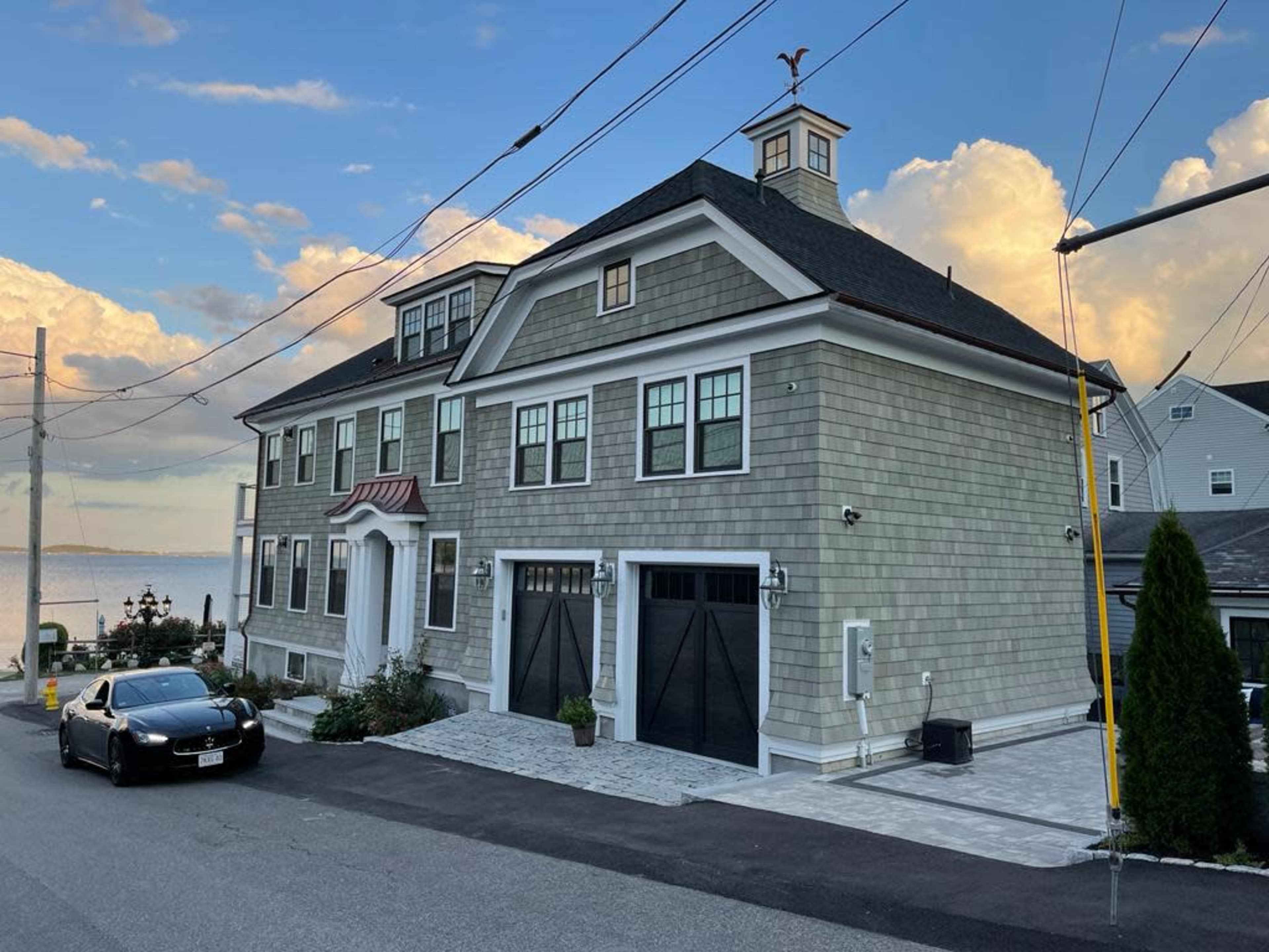 A large, multi-story shingle-style house with a prominent entrance and double garage doors overlooks a waterfront, with a car parked in the driveway.