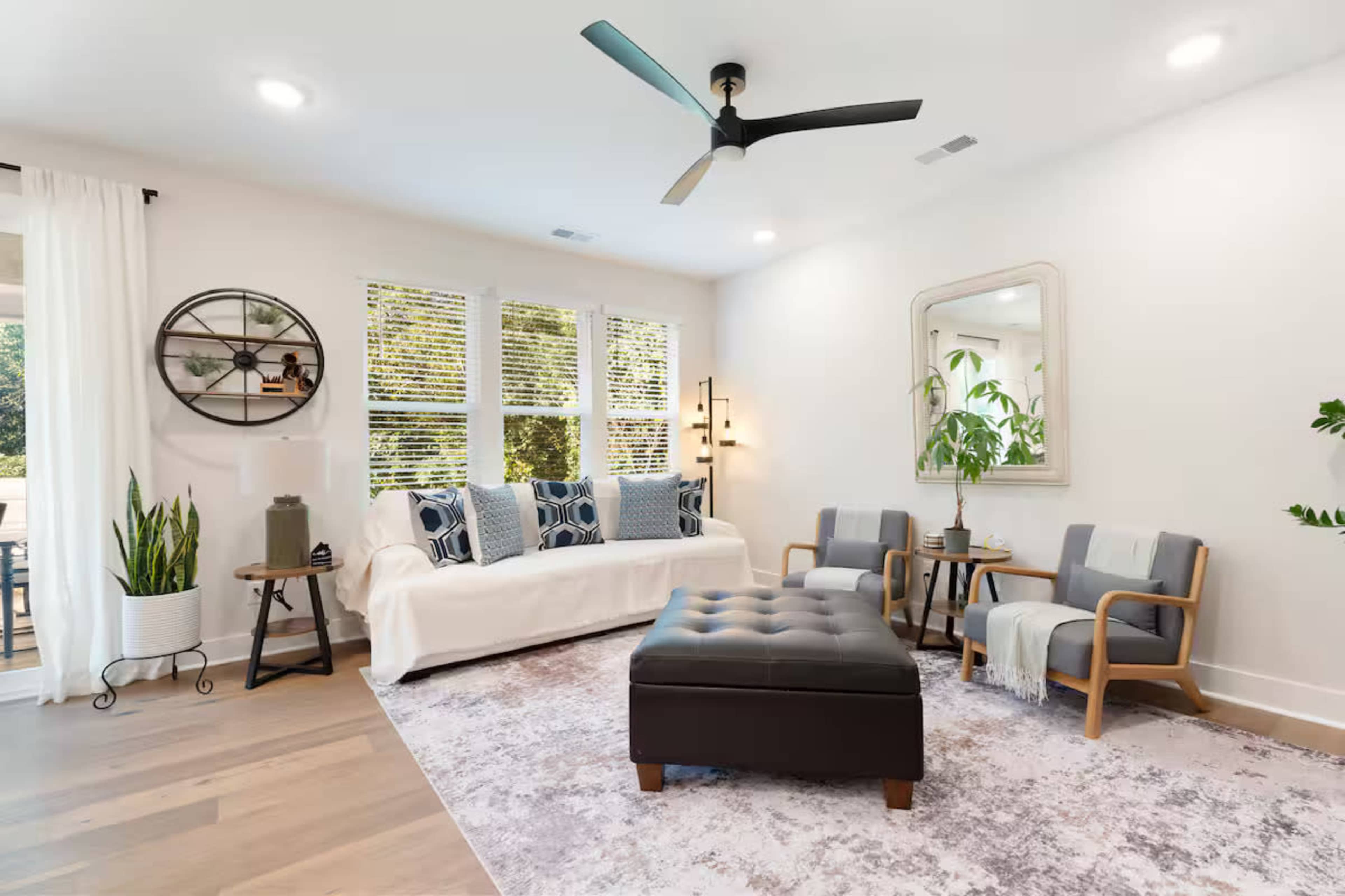 The image shows a modern living room featuring a white couch, a tufted ottoman, two wooden chairs, and large windows allowing natural light to enter.