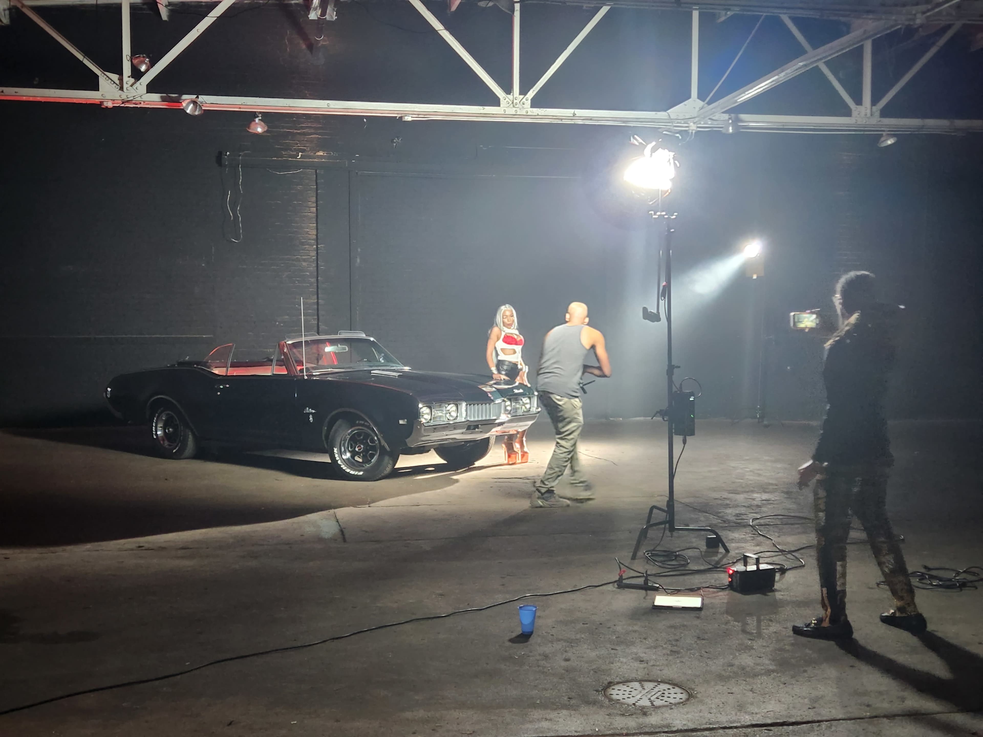 A classic black convertible car is positioned in a dimly lit industrial space while a person interacts with a model in front of it.