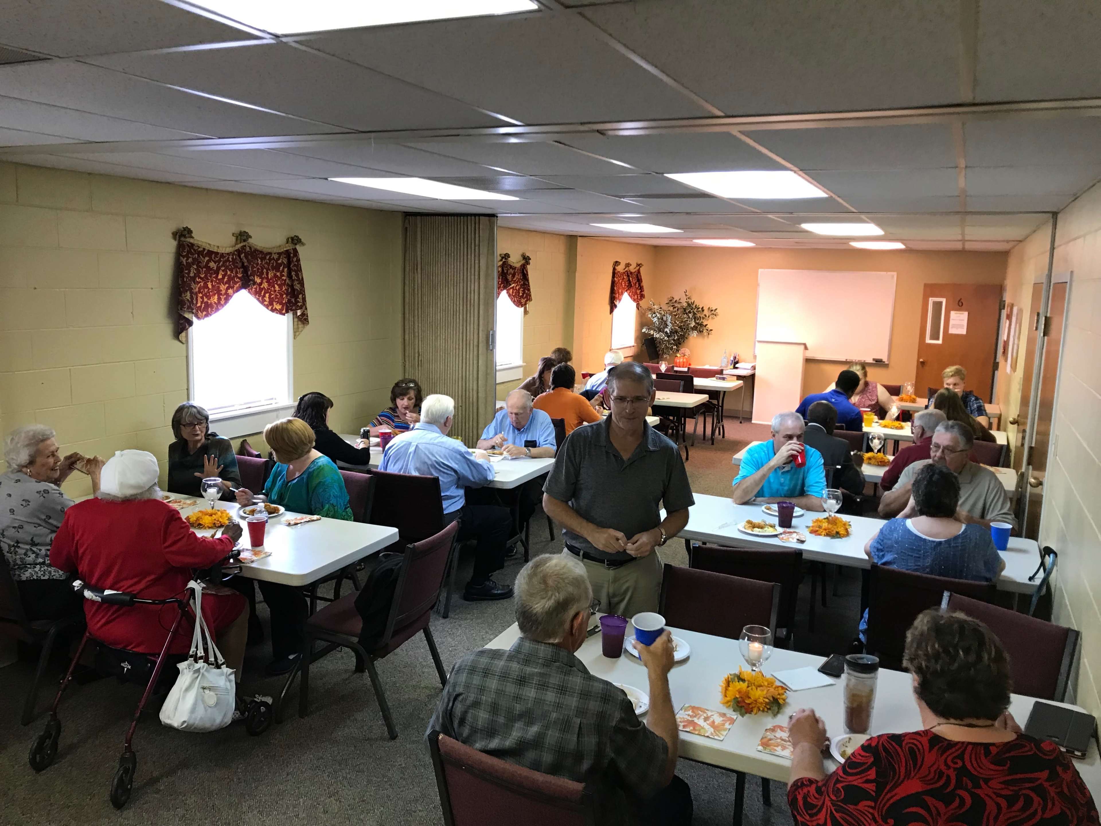 A group of people sits at tables in a community room, engaged in conversation and sharing a meal.