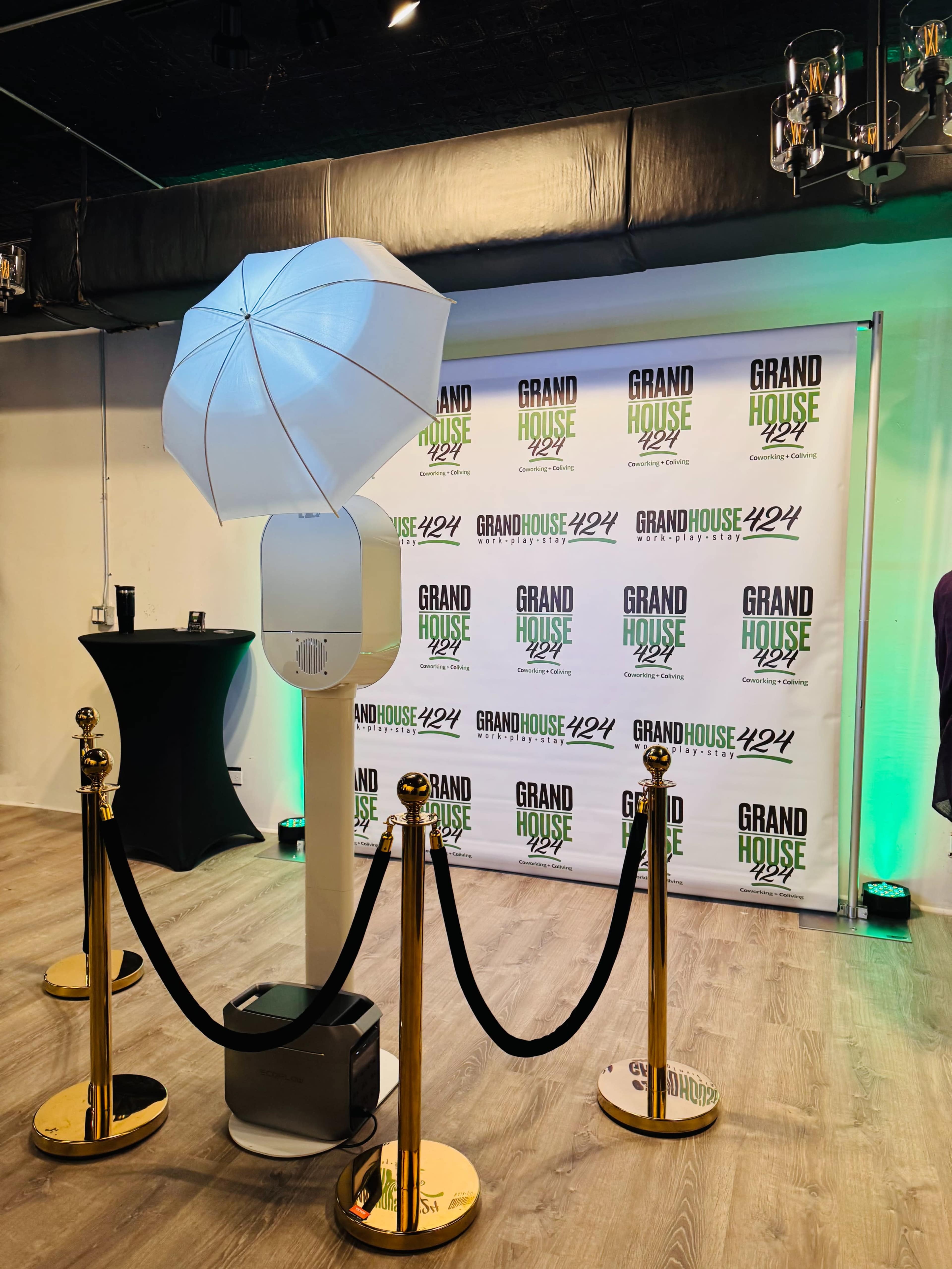 The image shows a photo booth with an umbrella light setup, surrounded by a red carpet barrier and a branded backdrop featuring the words "GRAND HOUSE 424" in multiple lines.