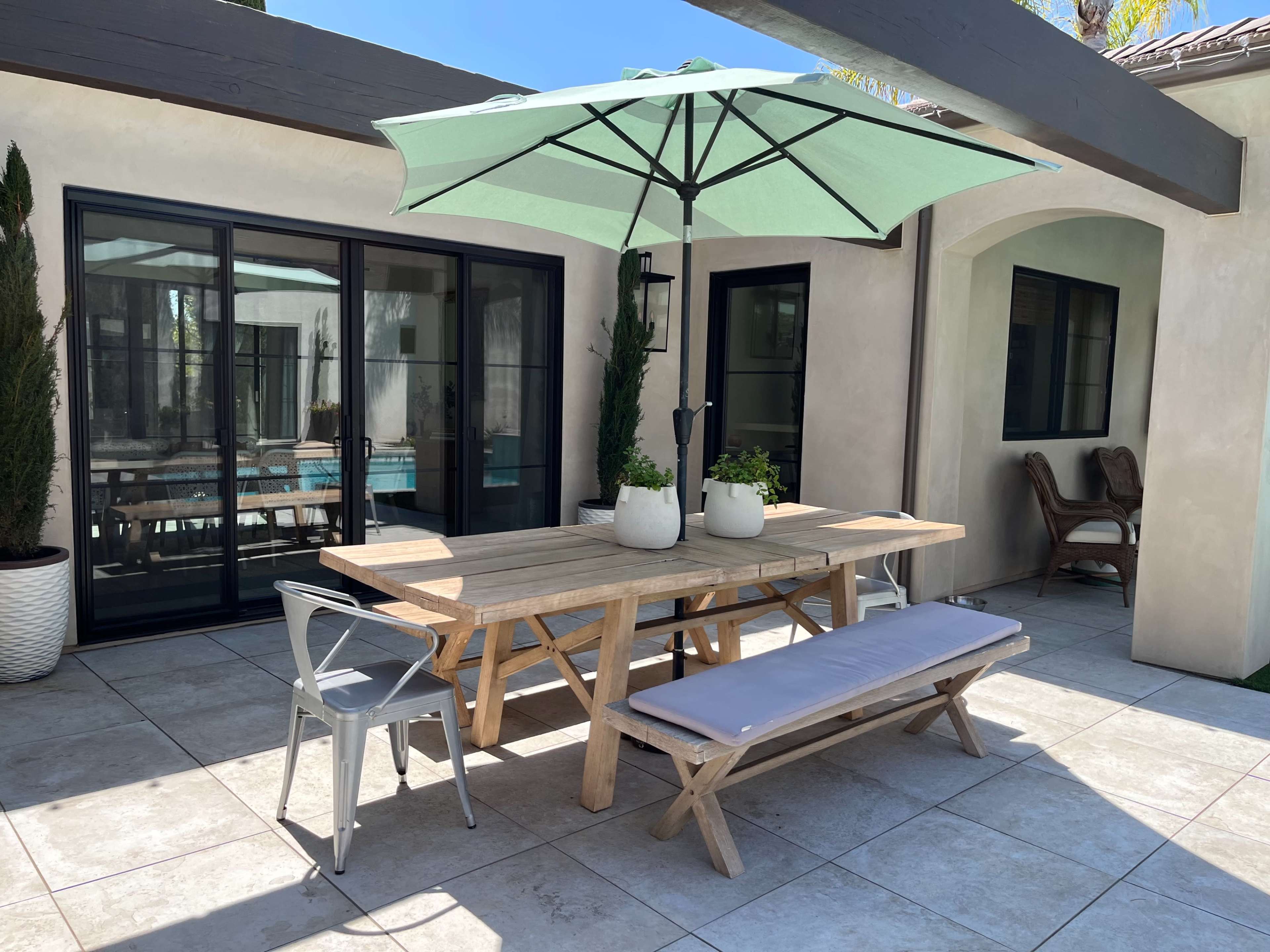 A wooden outdoor dining table with a green umbrella is set on a tiled patio adjacent to a house with large windows.