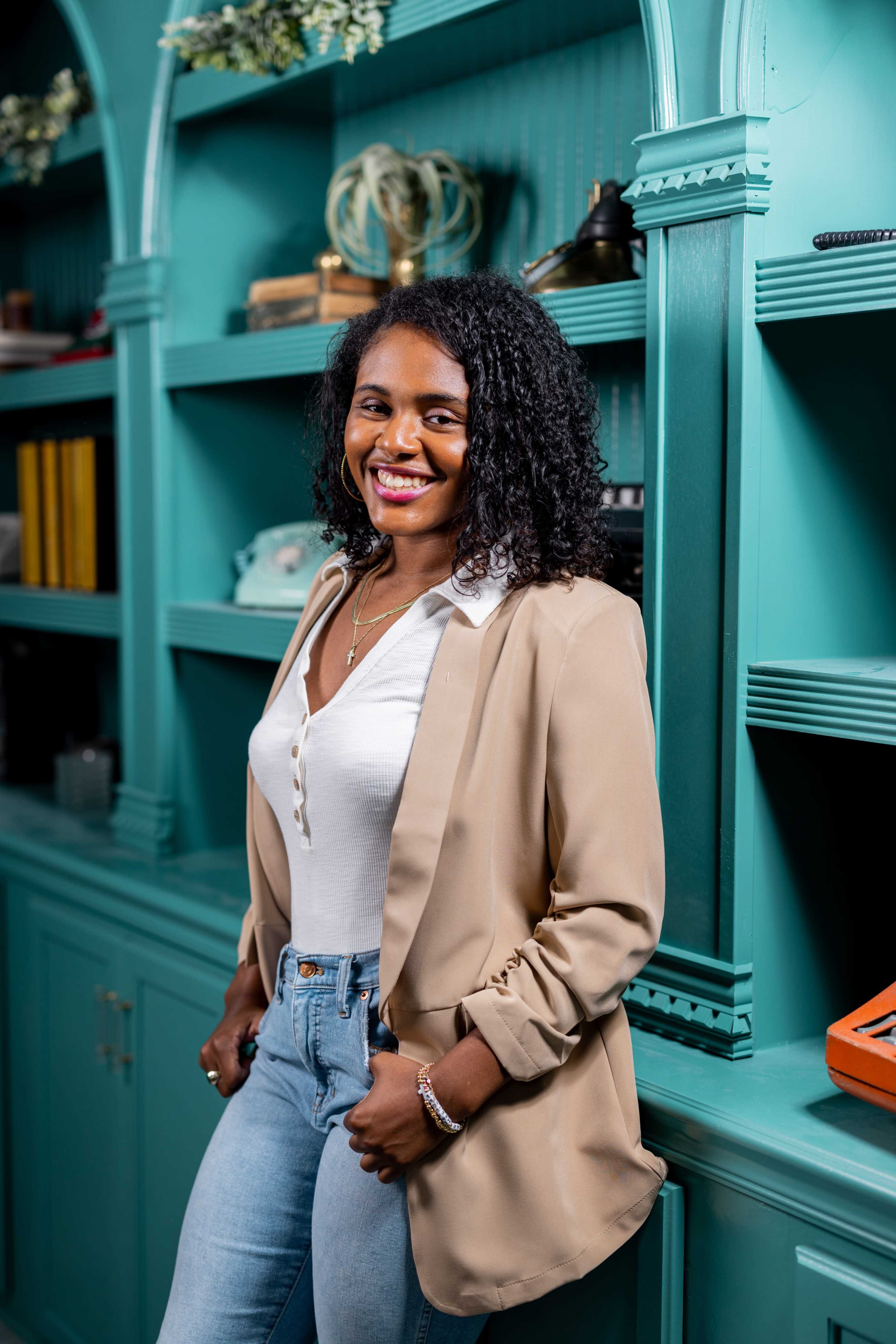 A woman with curly hair stands confidently next to a turquoise bookshelf, wearing a beige blazer and light jeans.