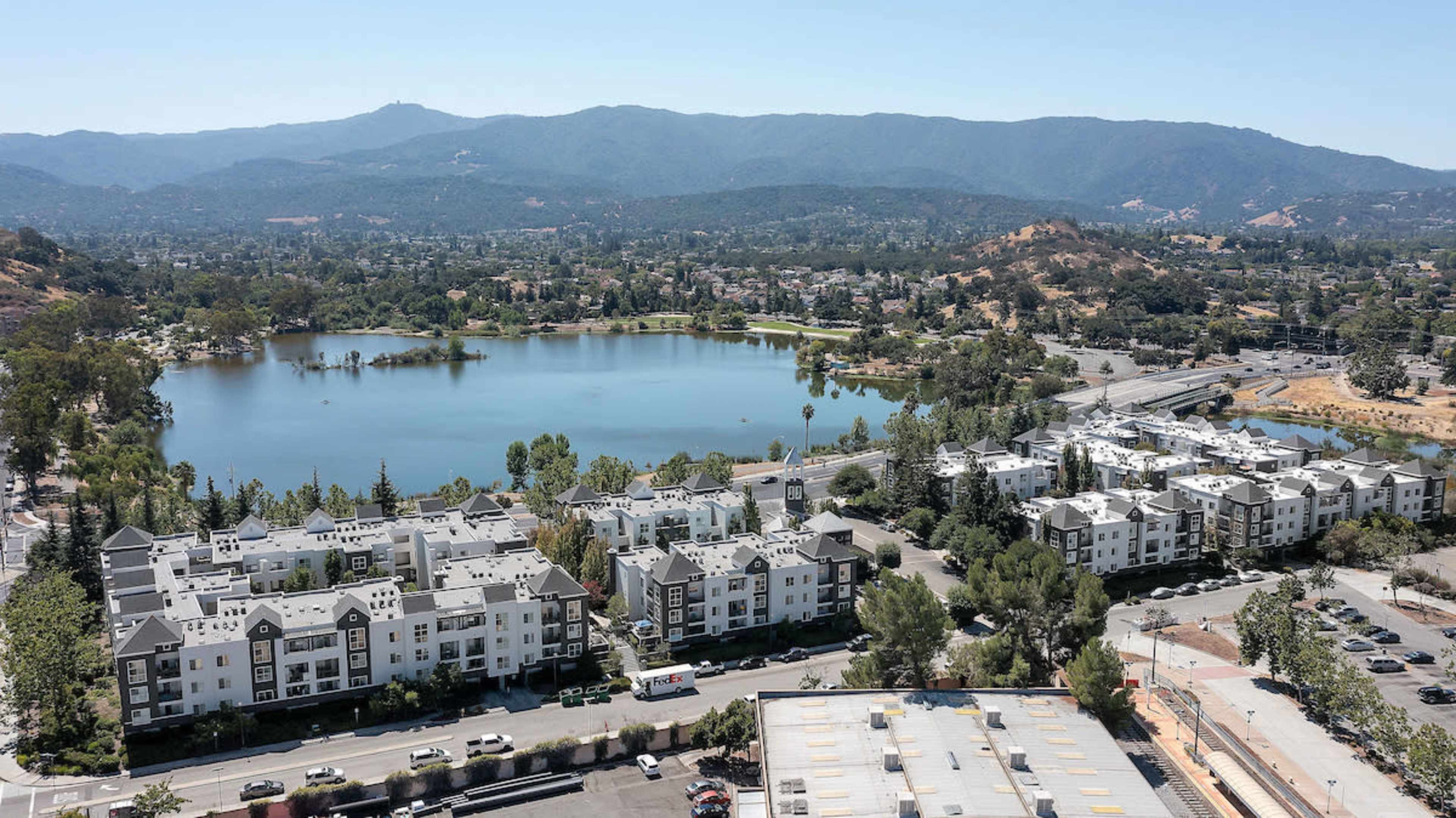 The image shows a serene lake surrounded by residential buildings and rolling hills in the background, with a highway running along the edge of the scene.