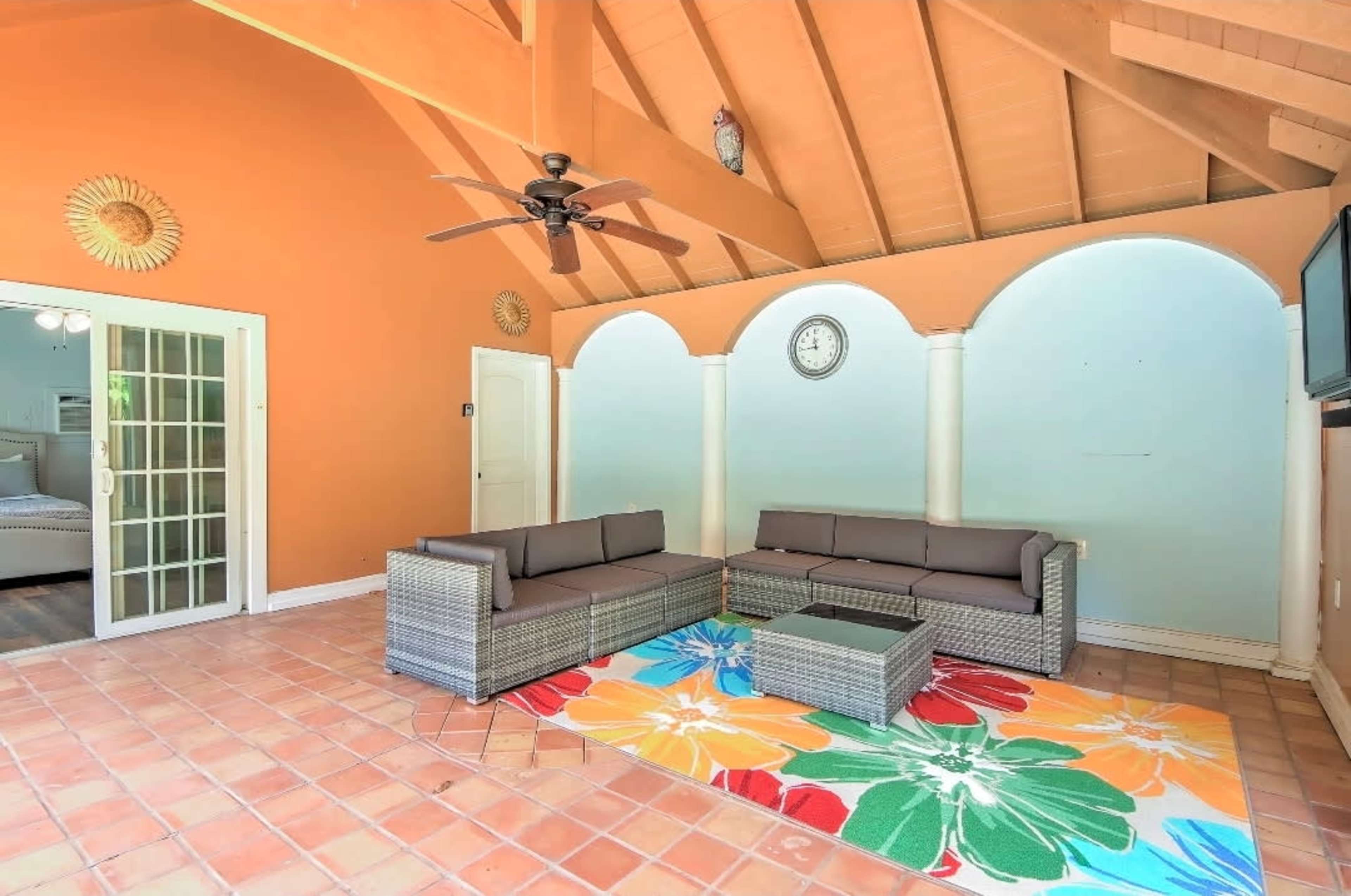 The image shows a spacious room with high wooden ceilings, featuring a seating area with a wicker sofa set, a colorful rug, and arched wall segments.