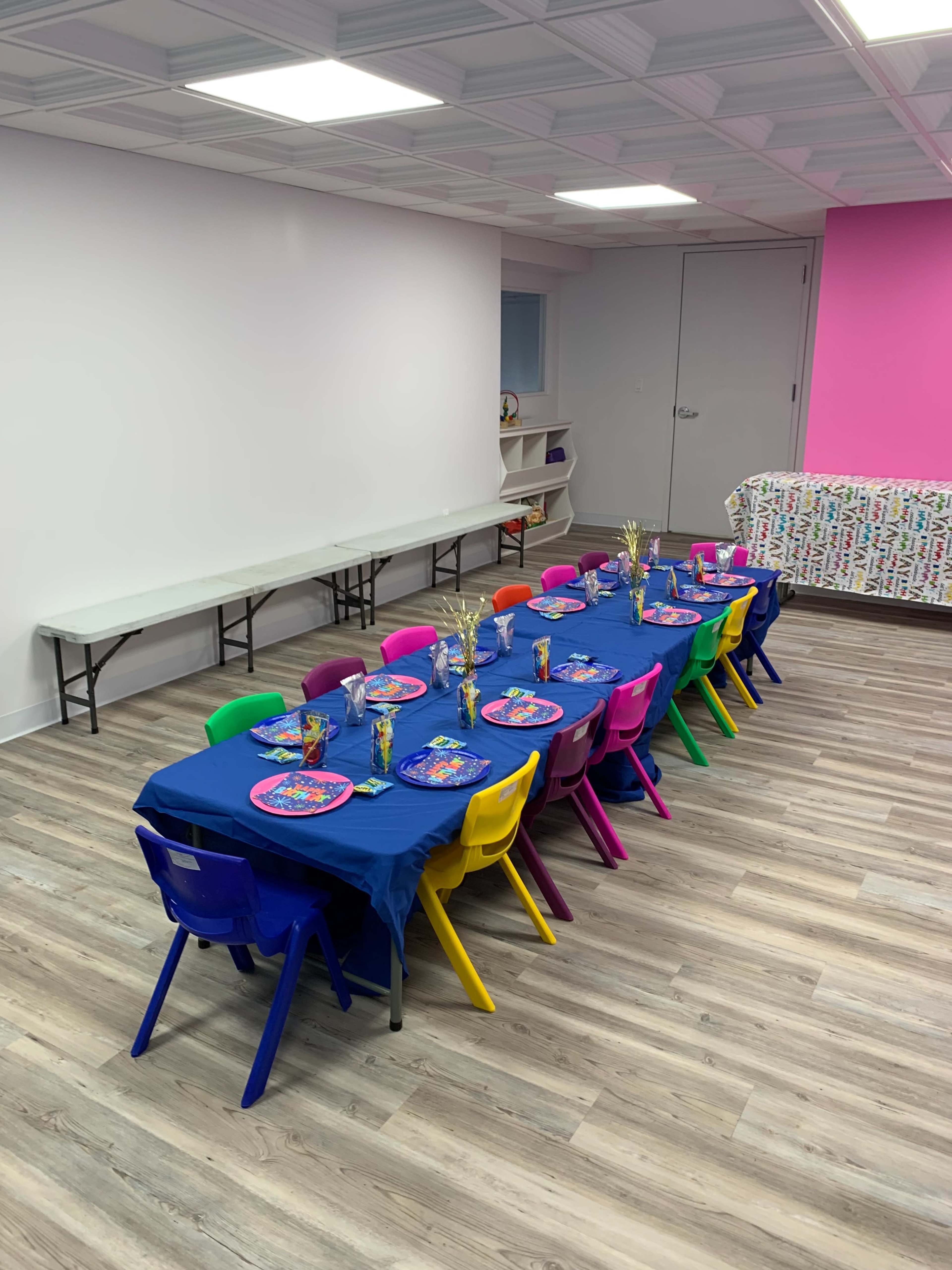 The image shows a long table set with colorful chairs, blue tablecloths, and decorative plates, in a brightly lit room with a pink wall.