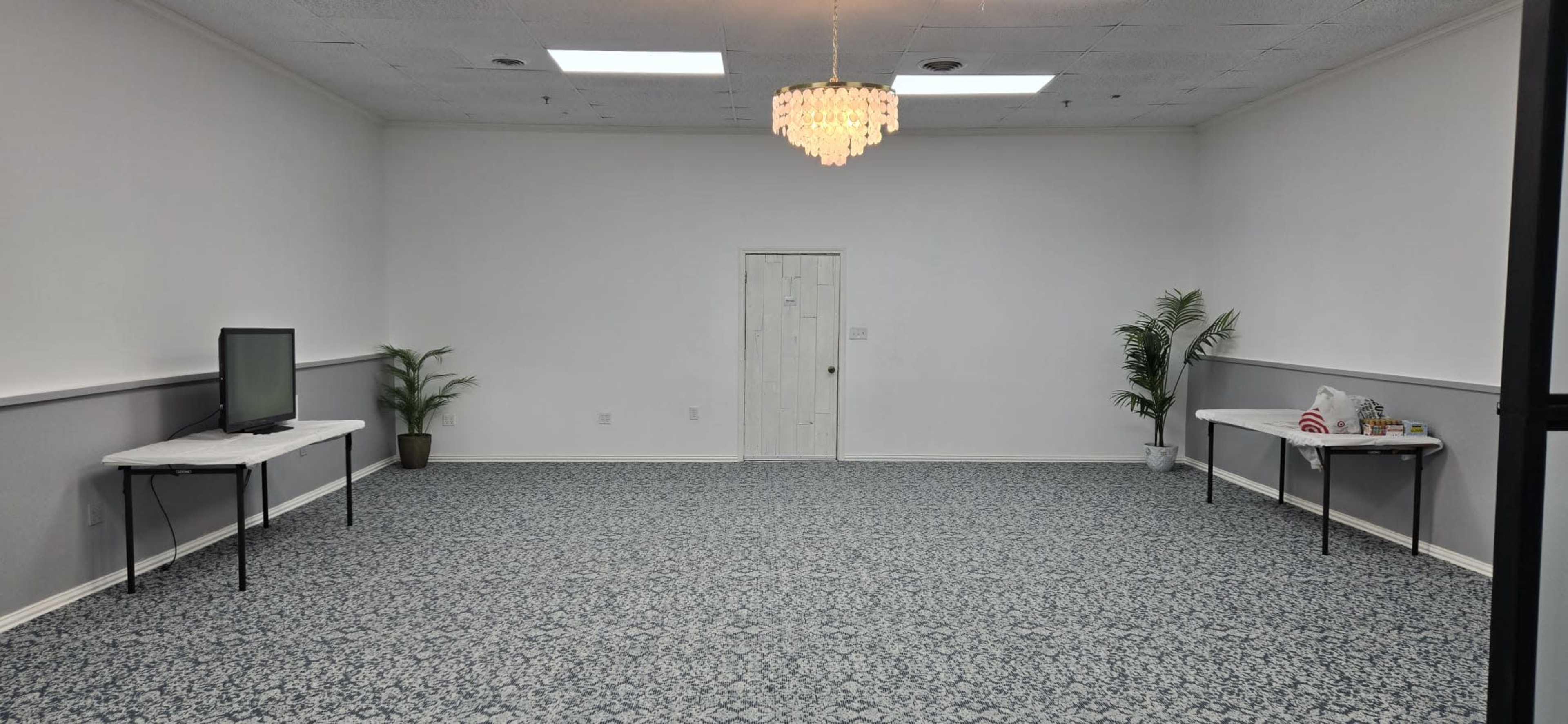 The image shows a spacious, empty room with white walls, a patterned carpet, a small chandelier, two tables with items on them, and a door in the center.
