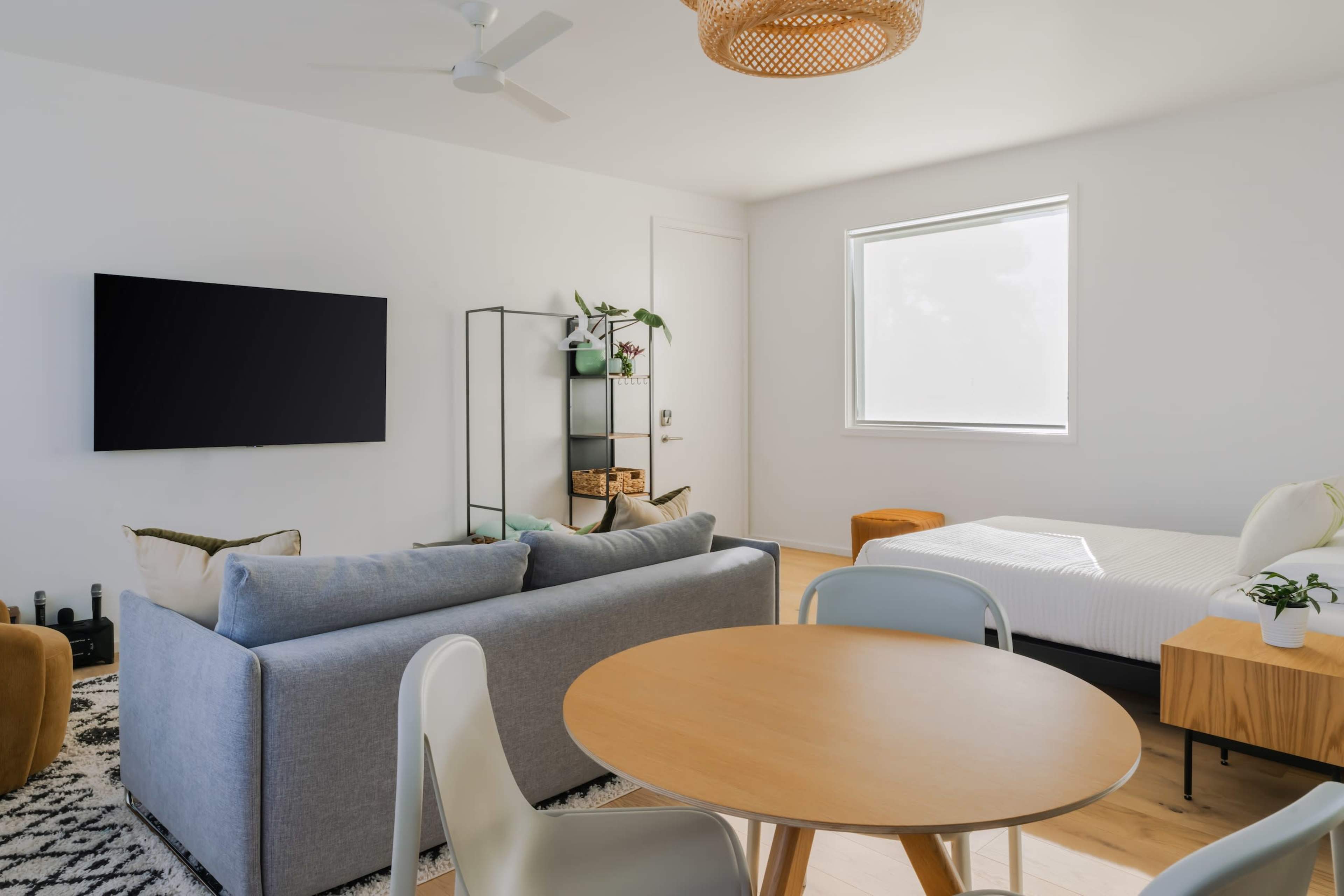A modern living space featuring a gray sofa, a circular wooden dining table, a bed, and a television mounted on the wall, with a small shelf displaying plants and decorative items.