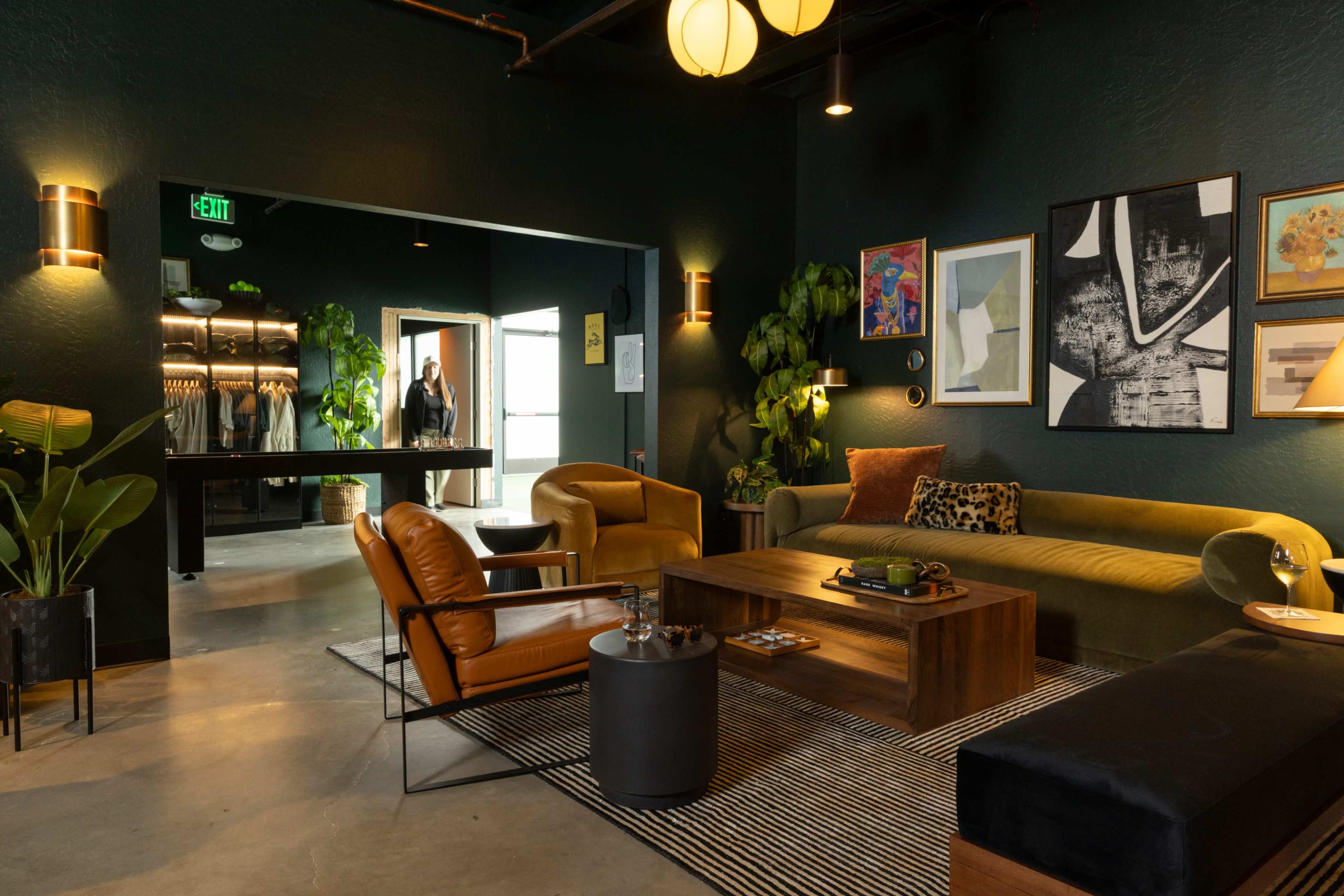 The image shows a stylish lounge area with a green wall, modern furniture, and plants, leading to an entrance with a coat rack in the background.